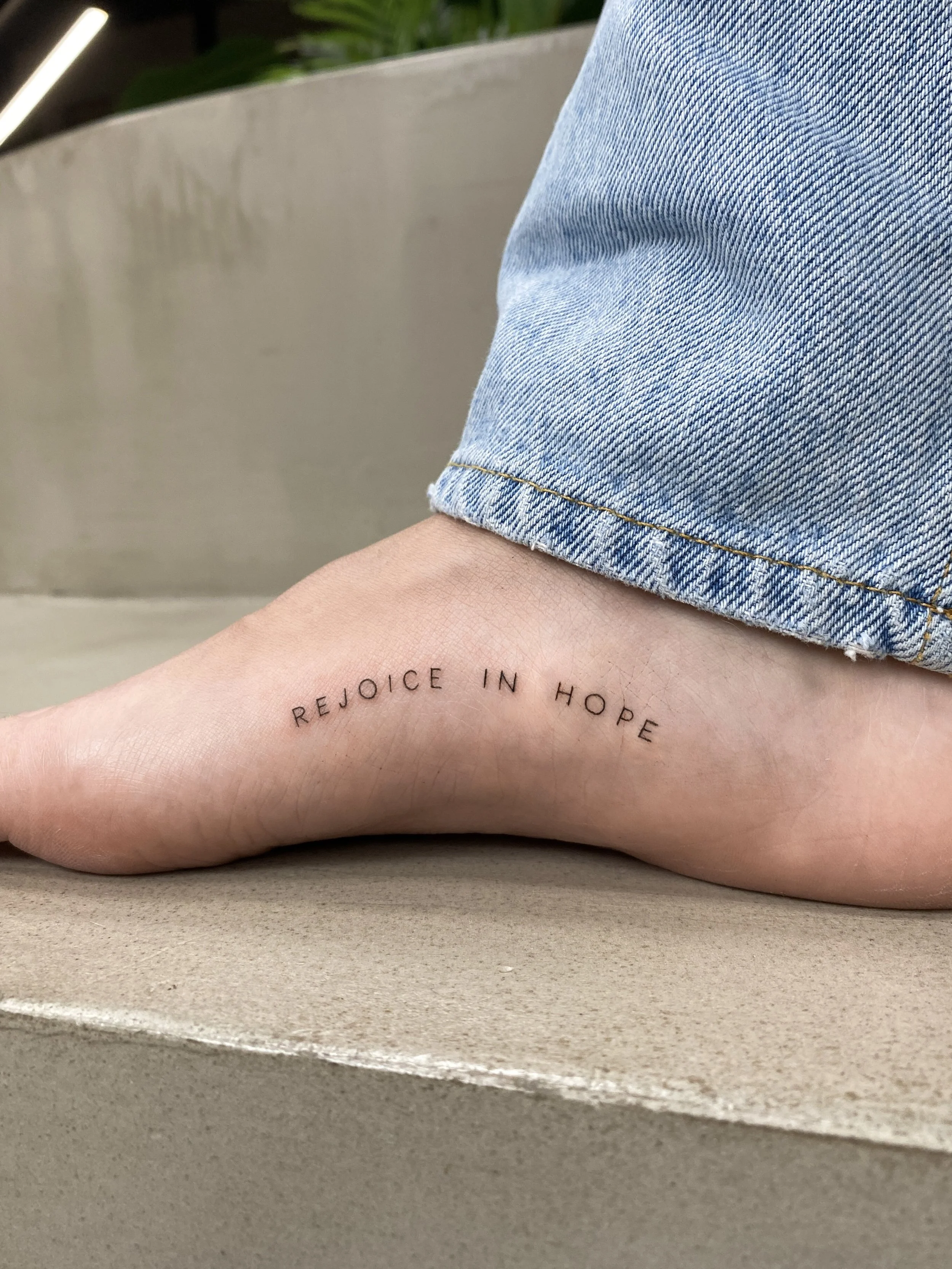 Tattoo on ankle reading 'Rejoice in Hope' with blue jeans and beige surface.