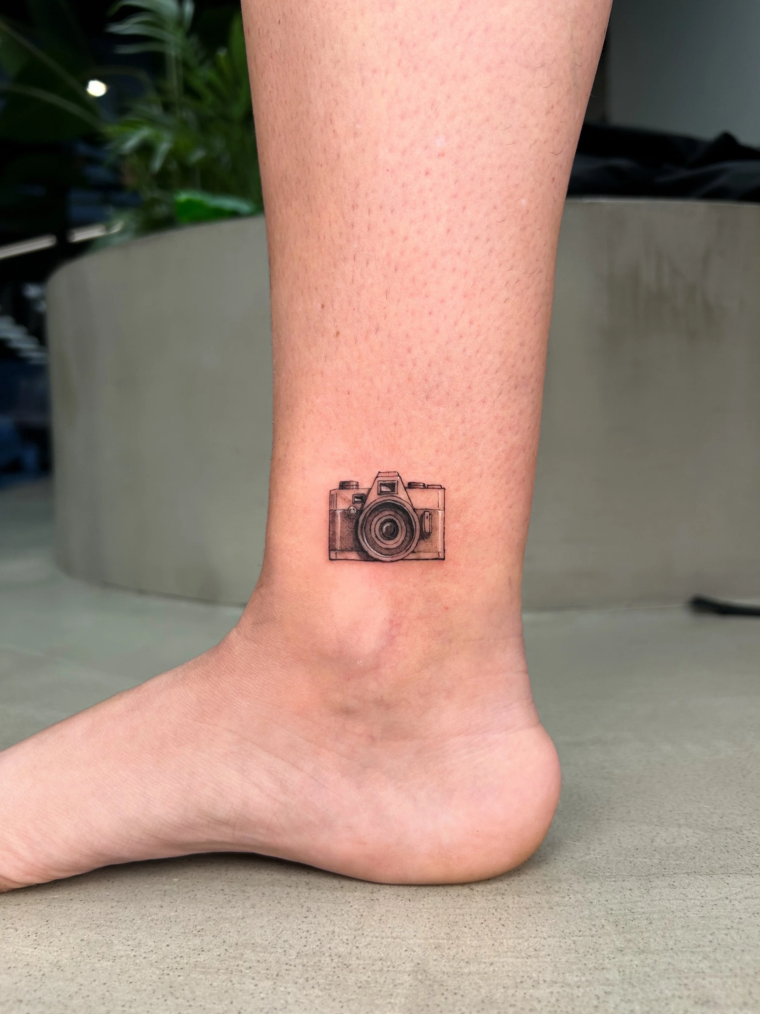Tattoo of a camera on the ankle of a person.