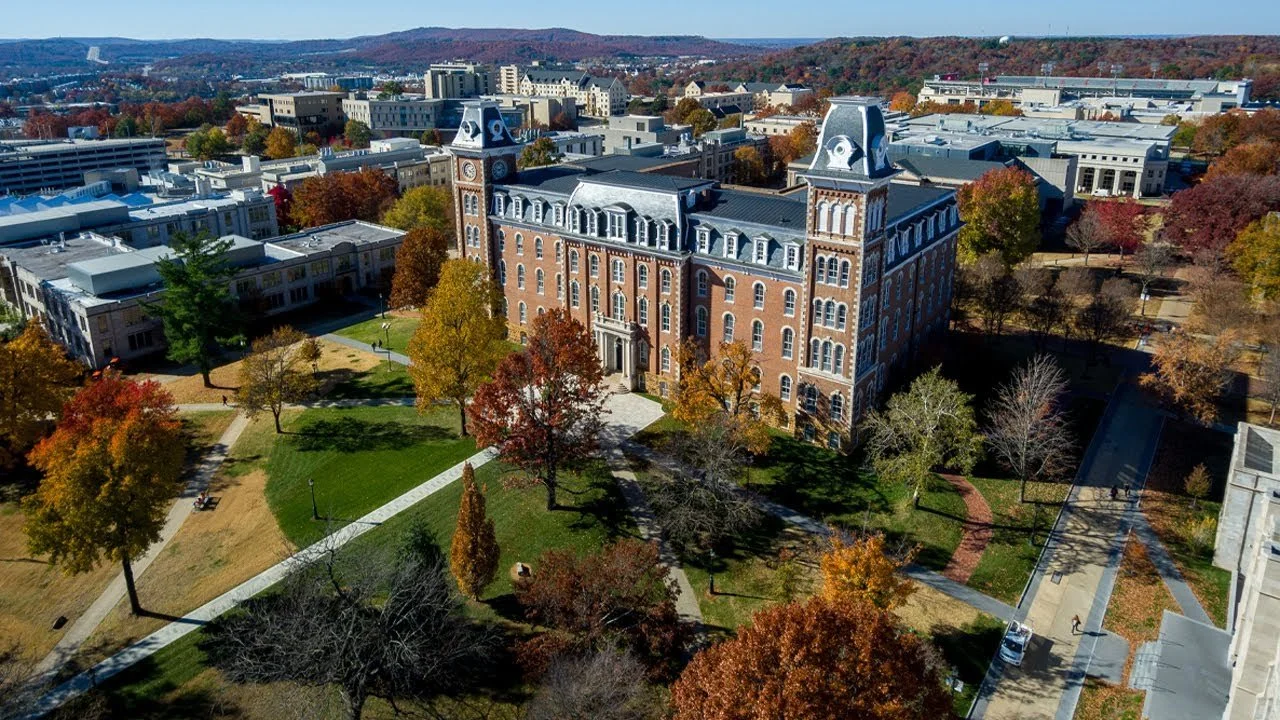 An aerial view of a historic brick building with clock towers on a college campus, surrounded by trees with fall foliage, walkways, and nearby modern buildings in the background.