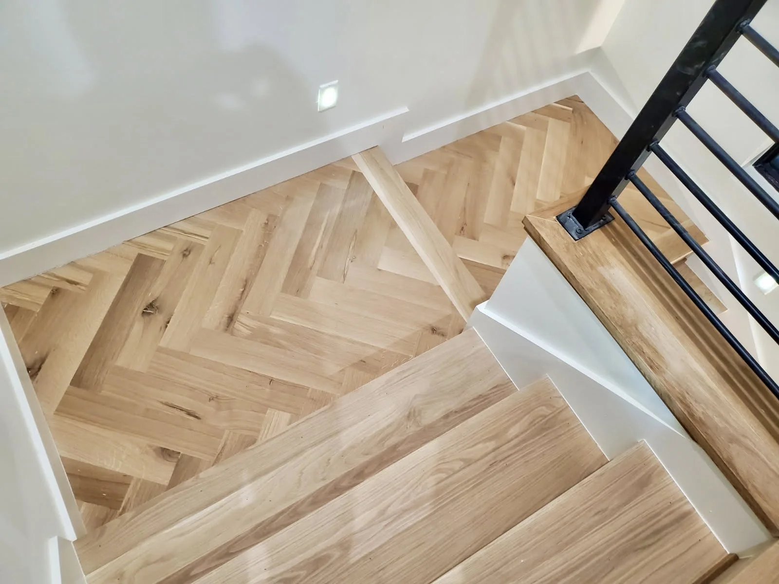 Top view of wooden staircase with black metal railing and herringbone patterned hardwood floor.