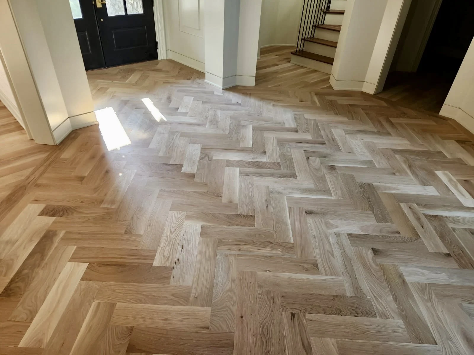 Interior view of a room with a herringbone wood floor, a black front door, a staircase with wooden steps and black metal railing, and an open doorway leading to a dark room.