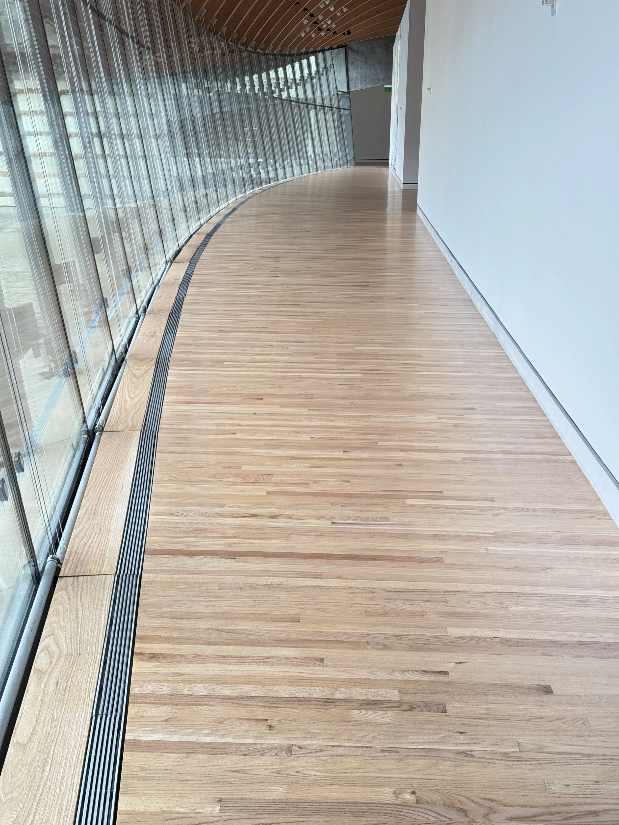 Curved wooden floor corridor with glass wall on the left and a white wall on the right, in a modern building.