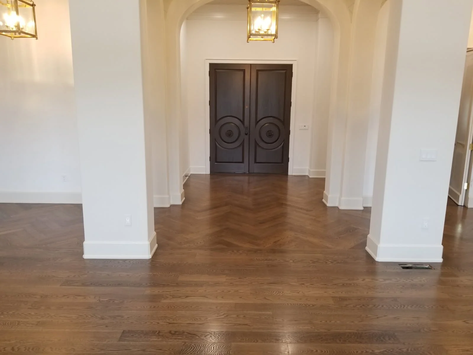 An empty room with hardwood floors, white walls, white trim, and a pair of dark wooden double doors at the far end. There are two hanging light fixtures and an air vent on the floor.