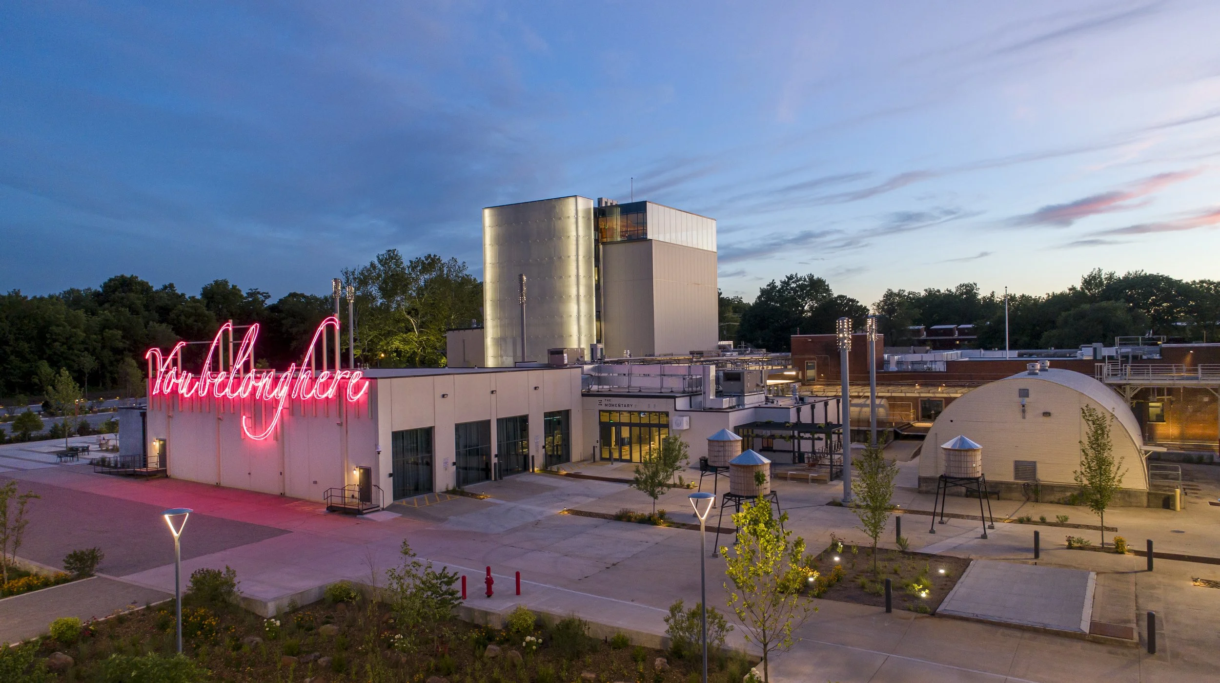 View of a modern building with a neon sign that says 'Yardlandhere' in pink lights, with trees in the background and a partly cloudy sky at dusk, outdoor lighting, small trees, and surrounding landscaping.