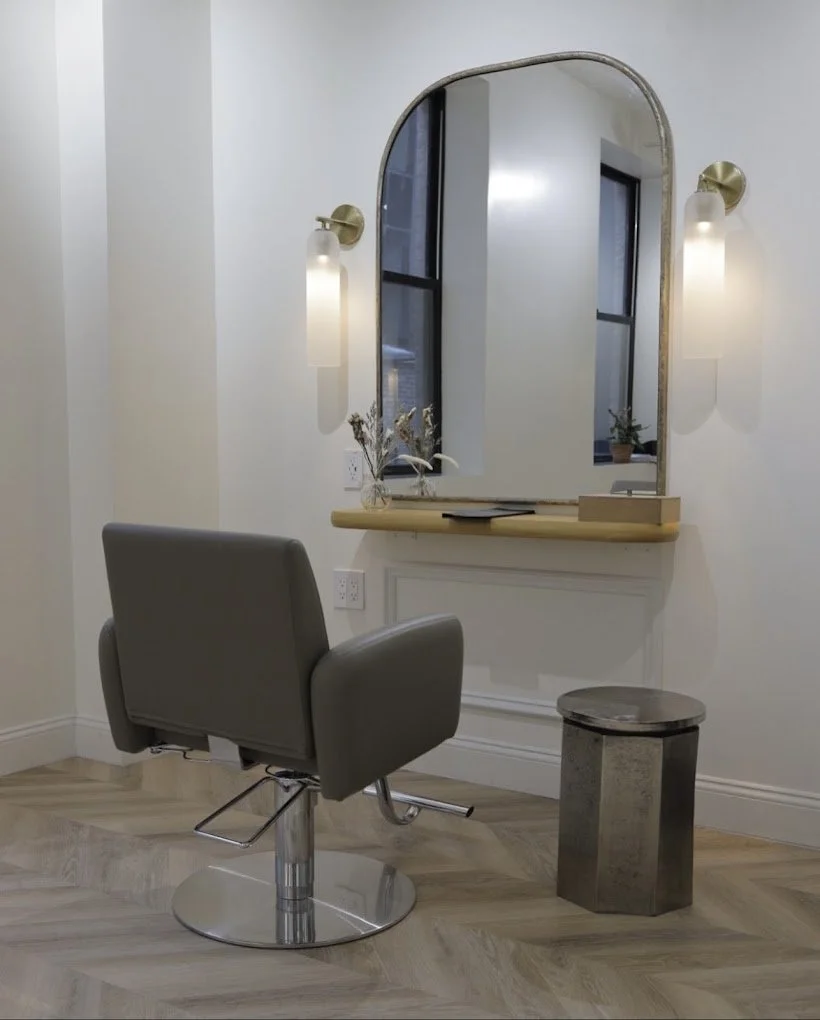 An empty salon chair facing a large mirror with wall-mounted lights on either side, next to a small metallic stool, a window, and a wooden shelf with decorative items.