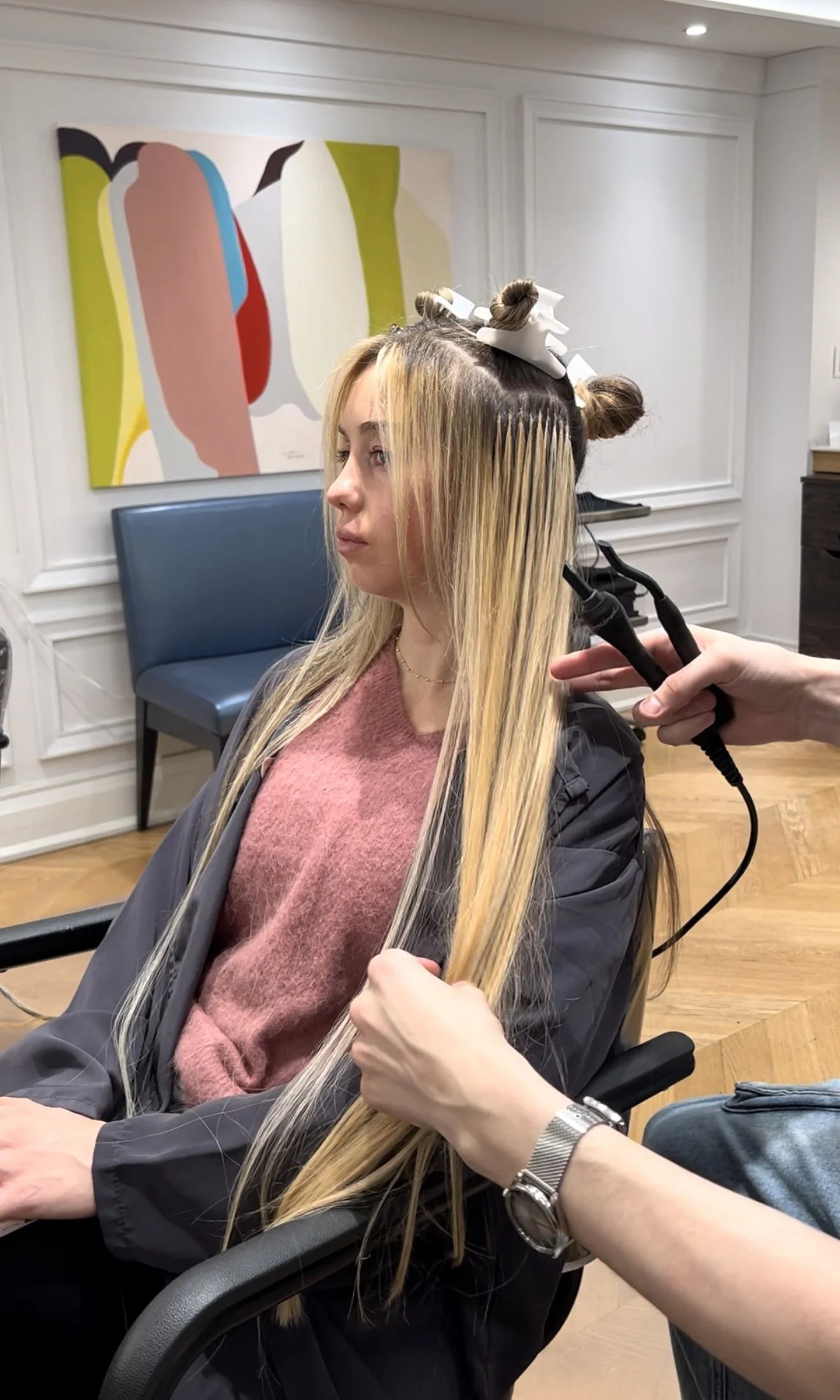 A woman with long blonde hair is sitting in a salon chair, getting her hair straightened with a flat iron. She has hair rollers on the top of her head, and a stylist is using the flat iron on the woman’s hair.