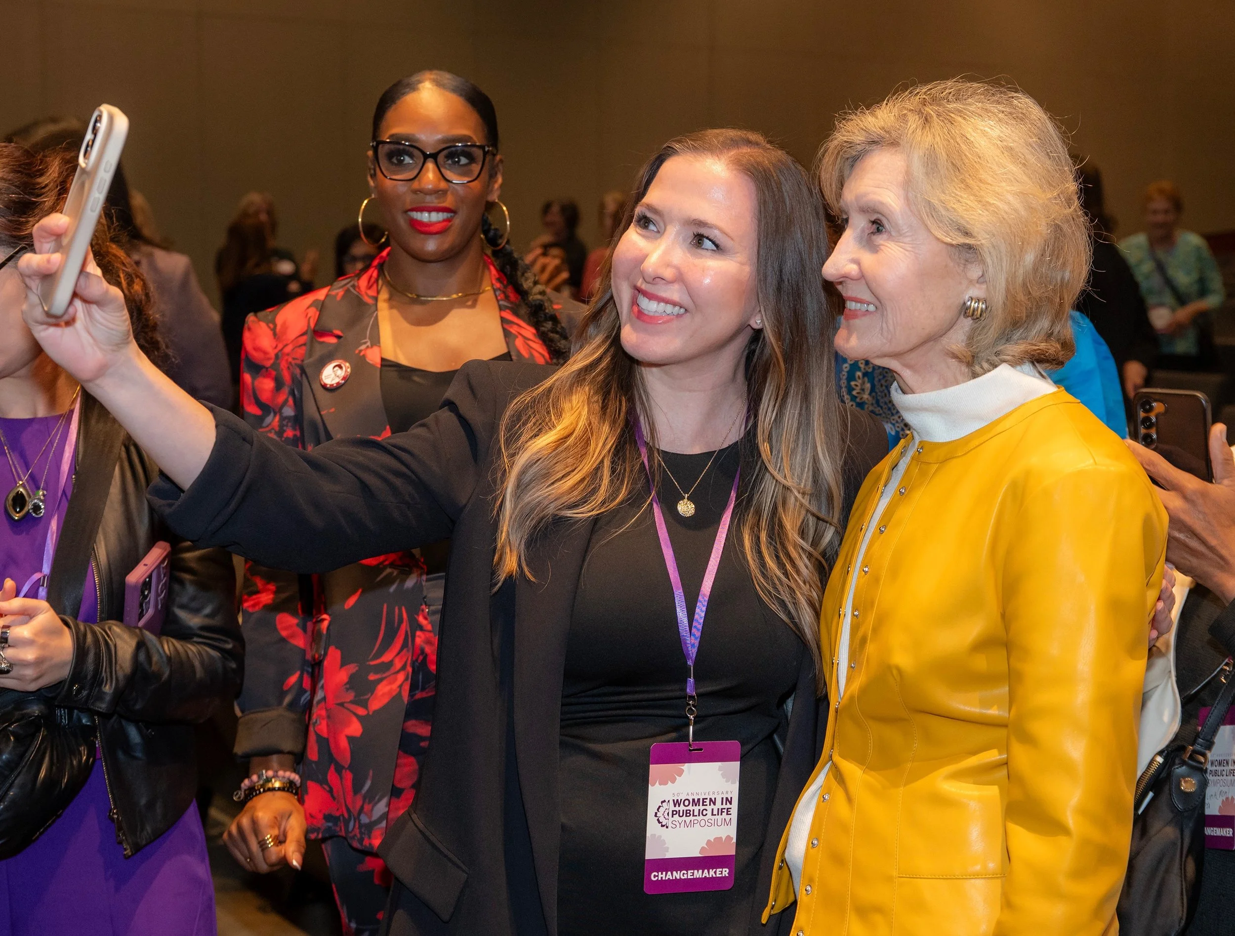 Three women taking a selfie at the Women in Public Life Symposium. One woman is holding a phone, and all are smiling and wearing name badges.
