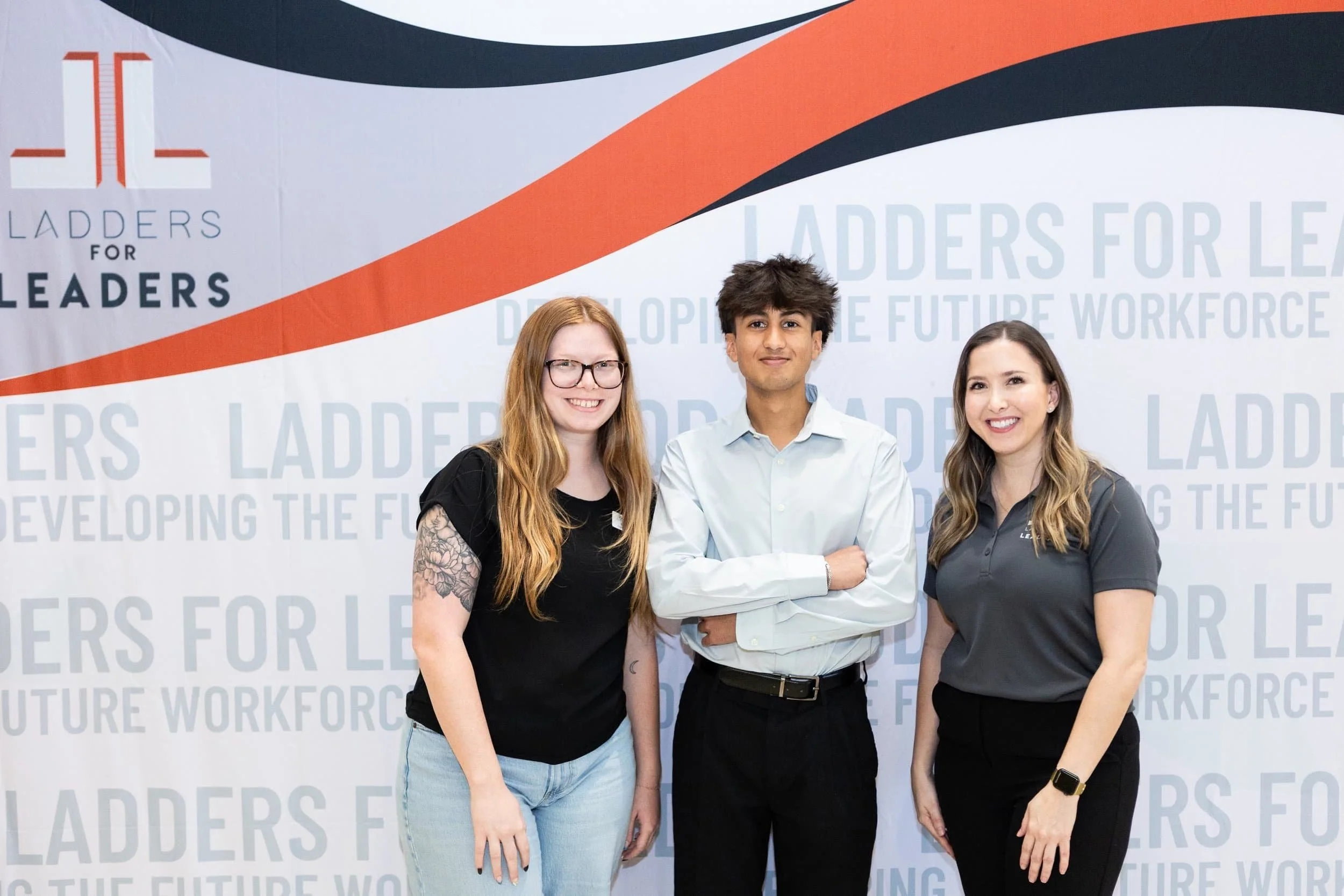 Three young people, two women and one man, standing and smiling in front of a backdrop that says "Ladders for Leaders" and "Developing the Future Workforce." The woman on the left has long red hair, glasses, a tattoo on her arm, and is wearing a blac
