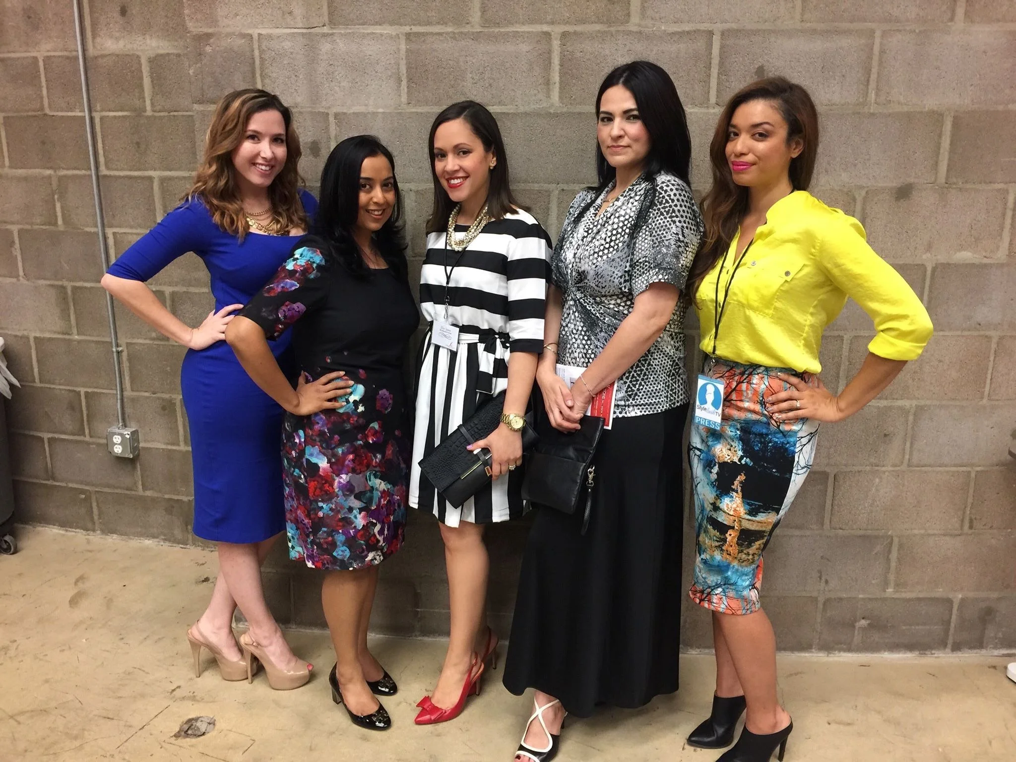 Group of five women standing against a brick wall, dressed in stylish clothing, posing for a photo.
