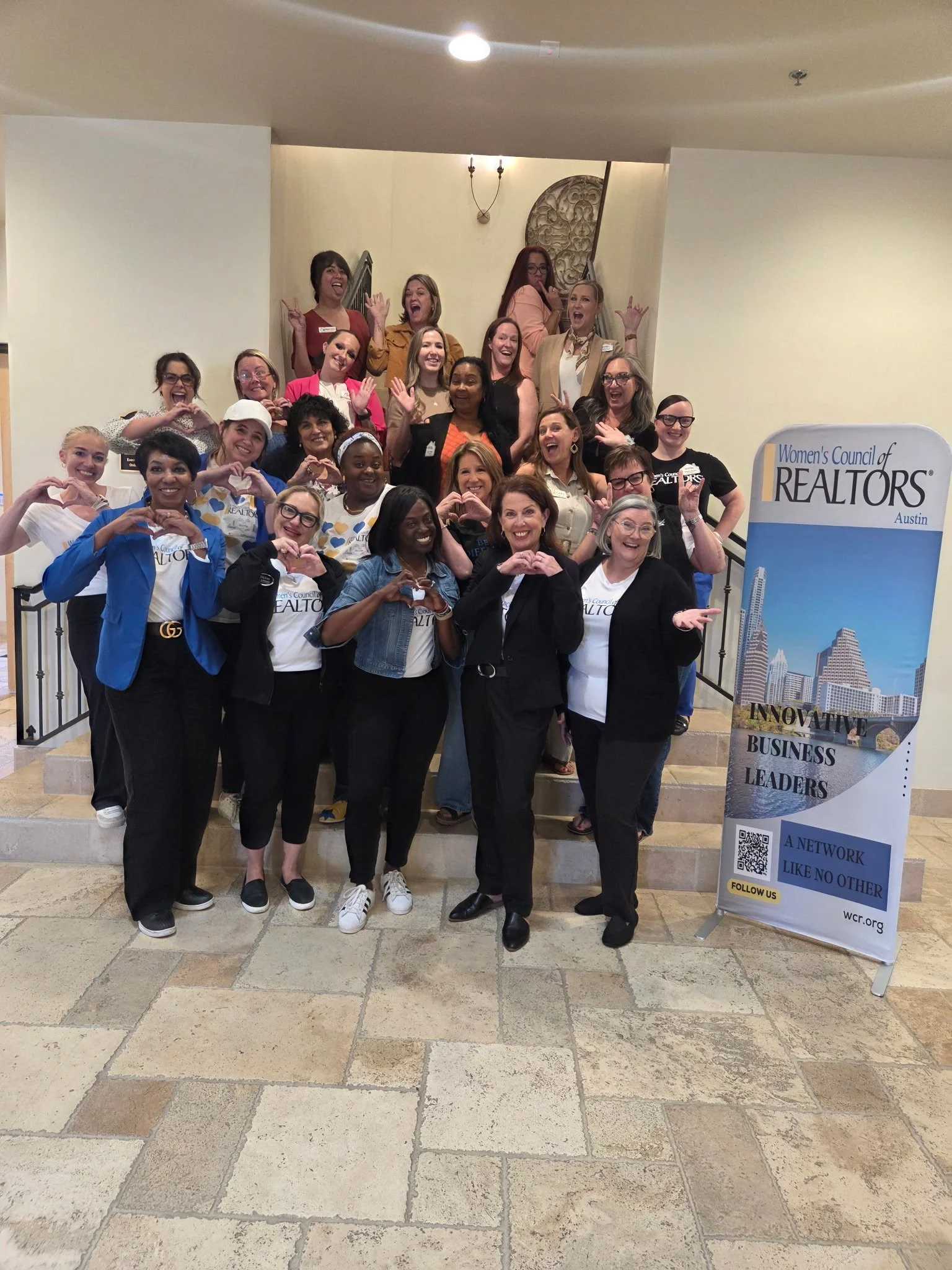 A group of women and men posing on a staircase at a Women's Council of Realtors event, making heart shapes with their hands, with a large vertical banner on the right side displaying the organization's logo and slogan.