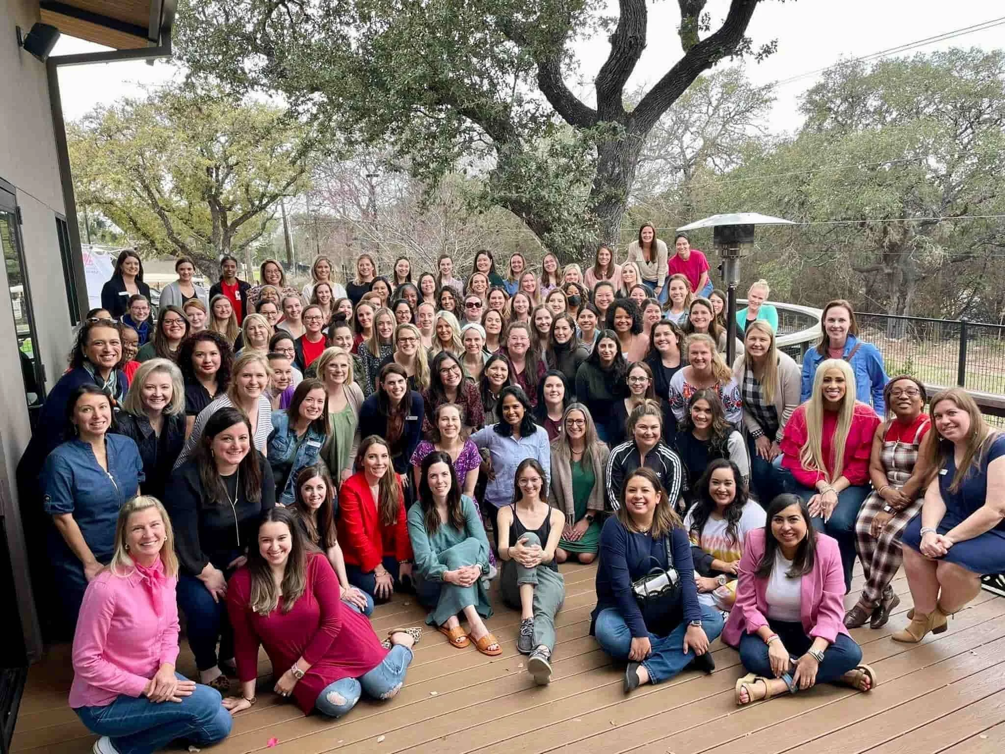 A large group of women posing for a photo outdoors on a wooden deck with trees in the background.