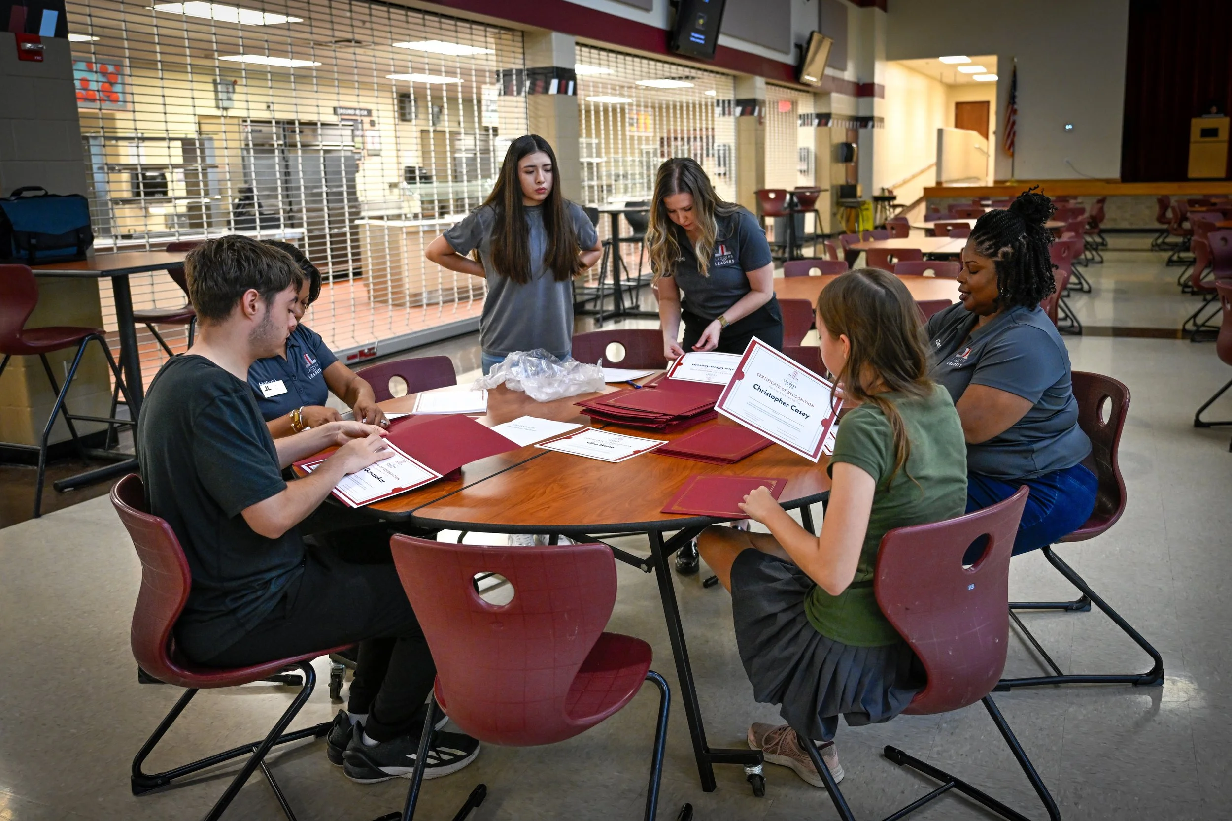 Group of six people sitting and standing around a table with red folders, preparing certificates in an indoor setting, possibly a school or community center.