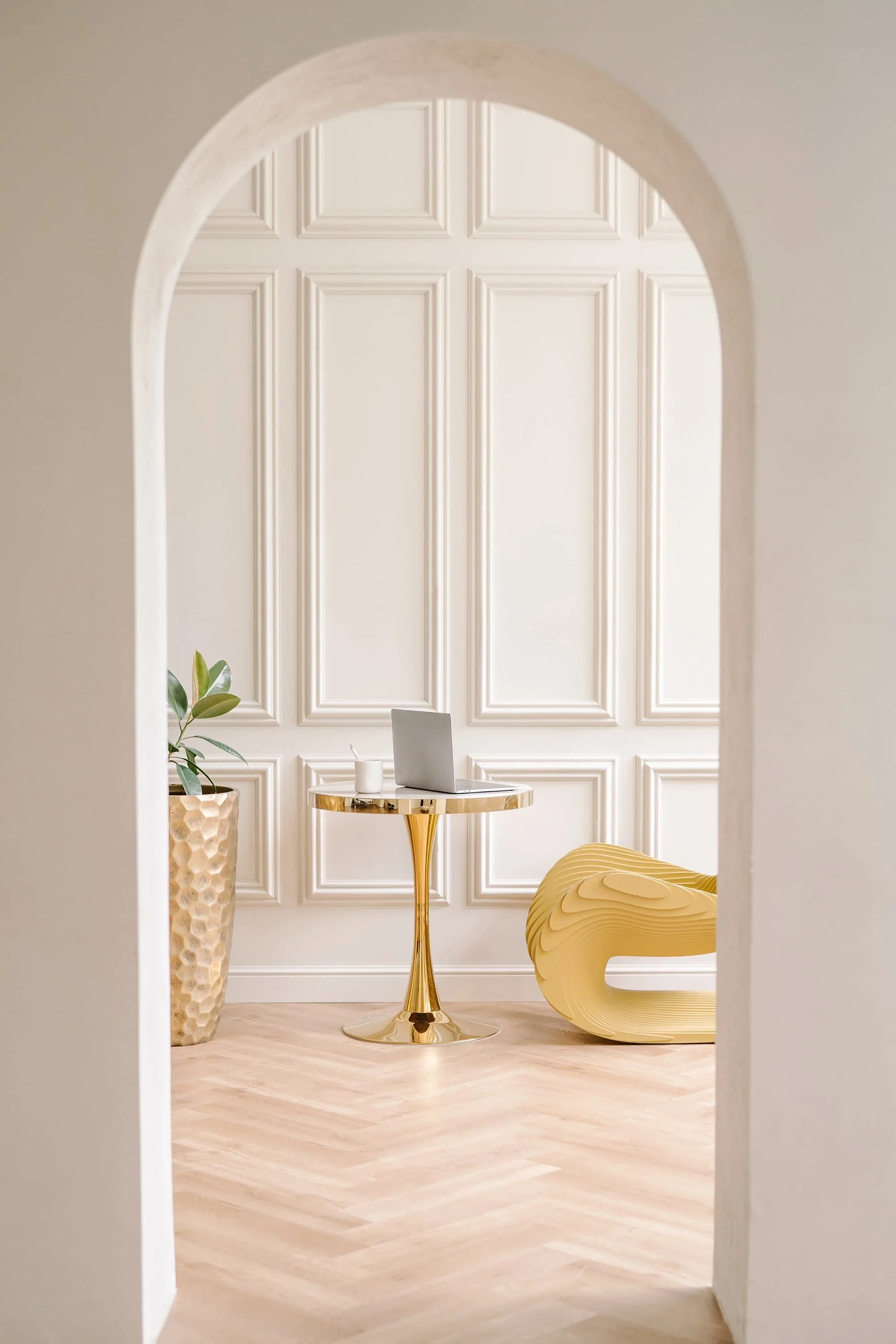 A modern interior space viewed through an arched doorway, featuring a small gold side table with a laptop, a white cup, and a mug. There is a textured gold vase with greenery on the left and a yellow sculptural chair on the right. The room has white paneled walls and light wood flooring.