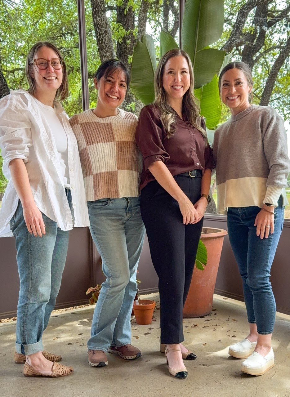 Four women smiling in a room with large windows and green trees outside, standing close together, with a large green plant behind them.