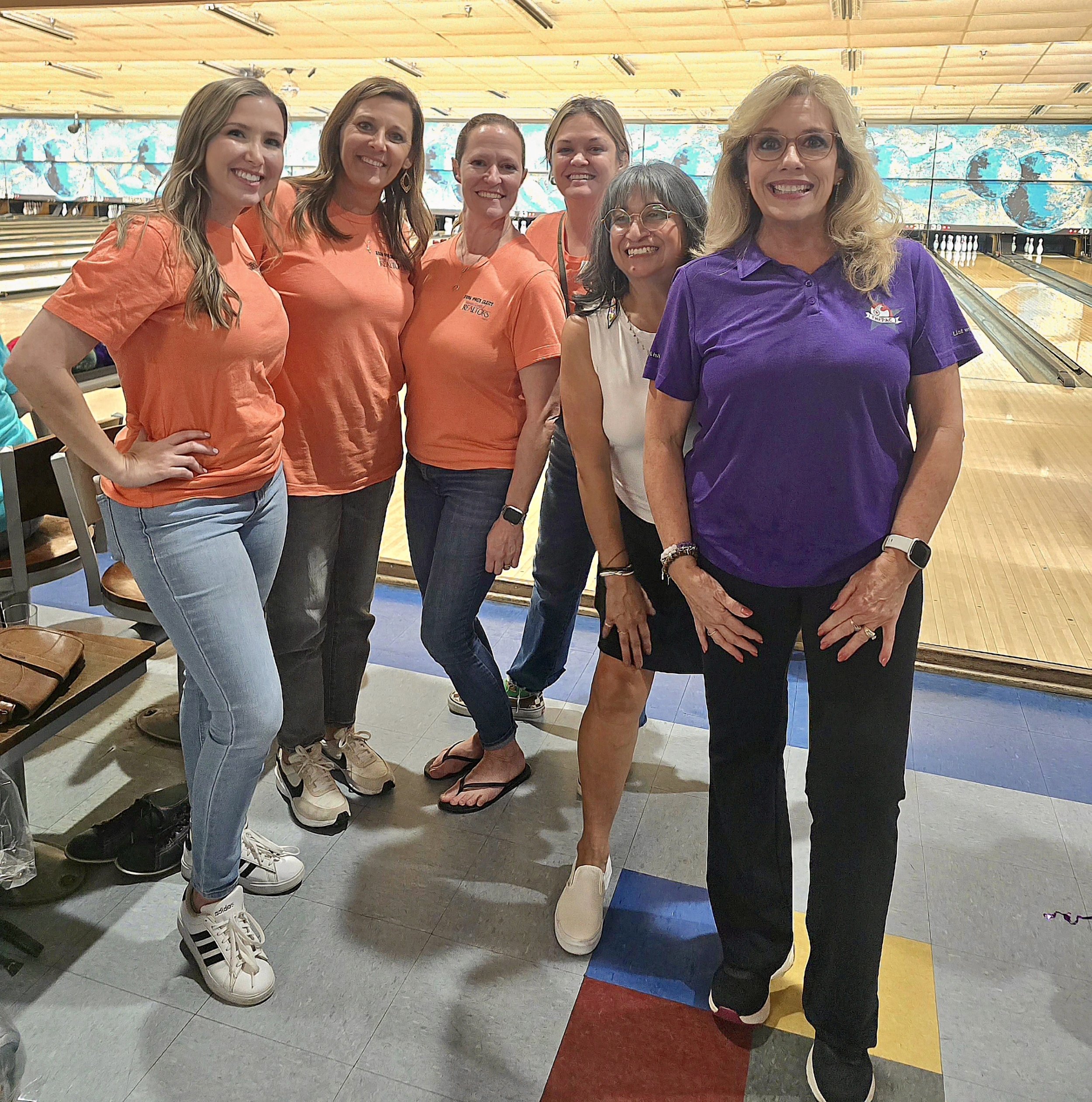 Group of six women posing for a photo at a bowling alley, with colorful mural artwork in the background.