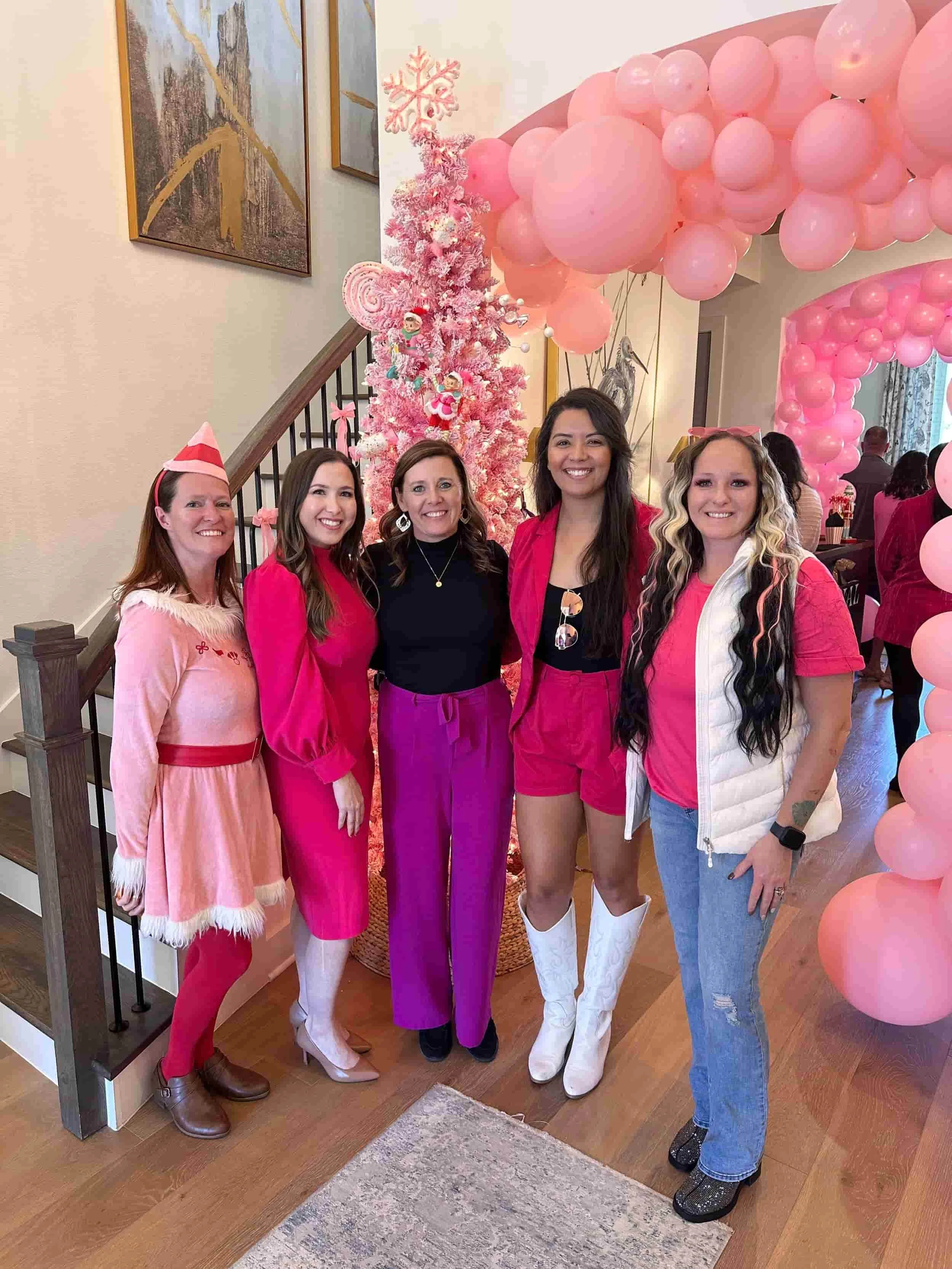 A group of five women dressed in pink and winter-themed attire standing in front of a Christmas tree decorated with pink ornaments, surrounded by pink balloon arches at a festive celebration.