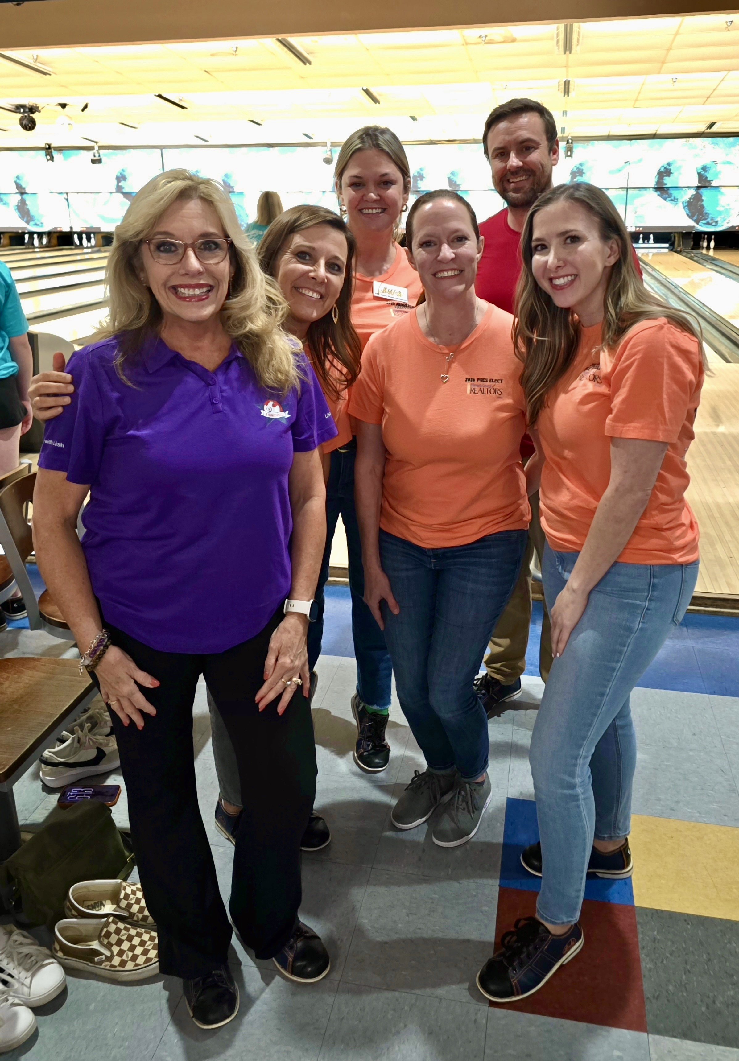 Group of six people at a bowling alley, smiling at the camera, with bowling lanes and screens in the background.