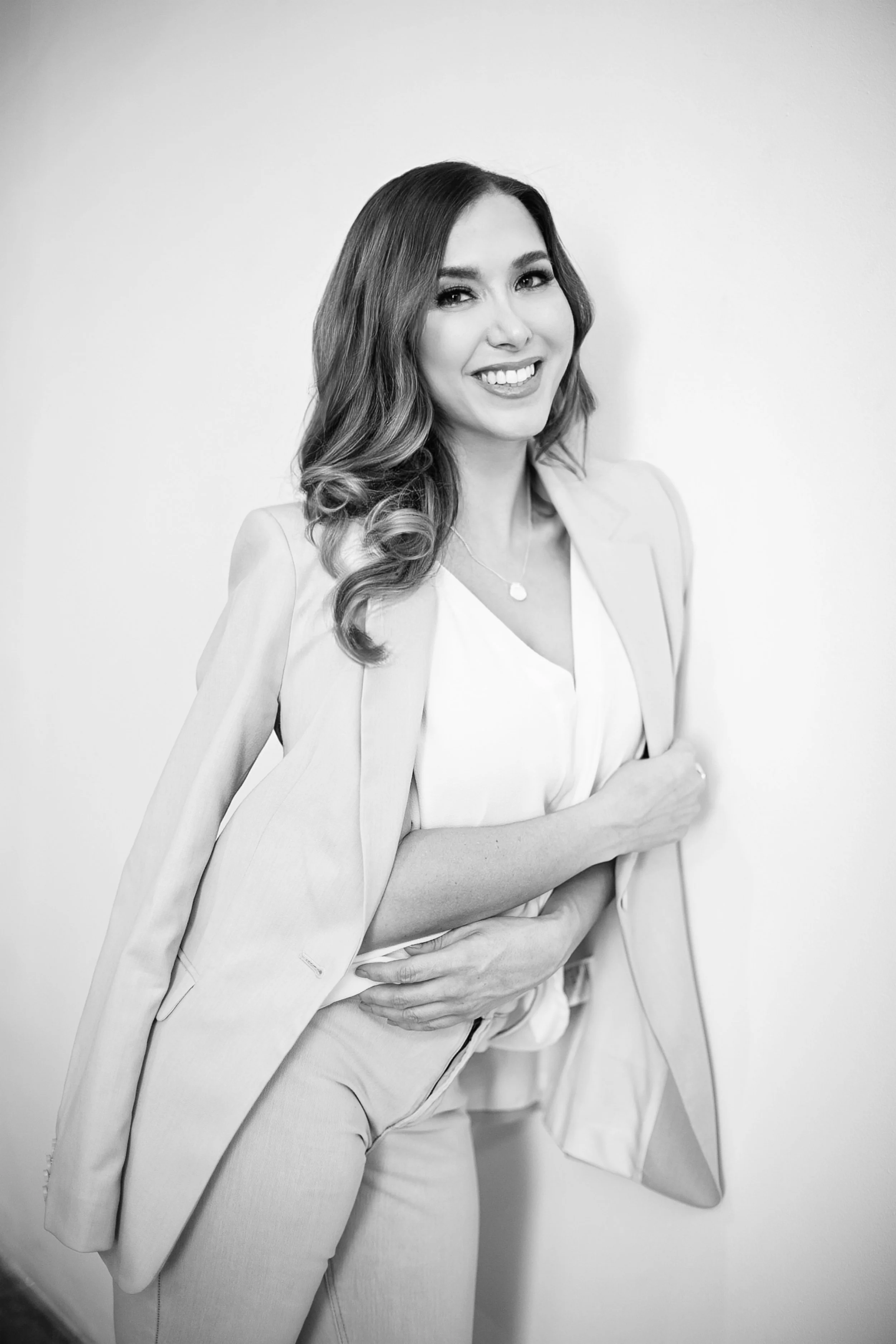 Black and white portrait of a woman smiling, casual pose, wearing a blazer and blouse, with wavy hair, standing against plain wall
