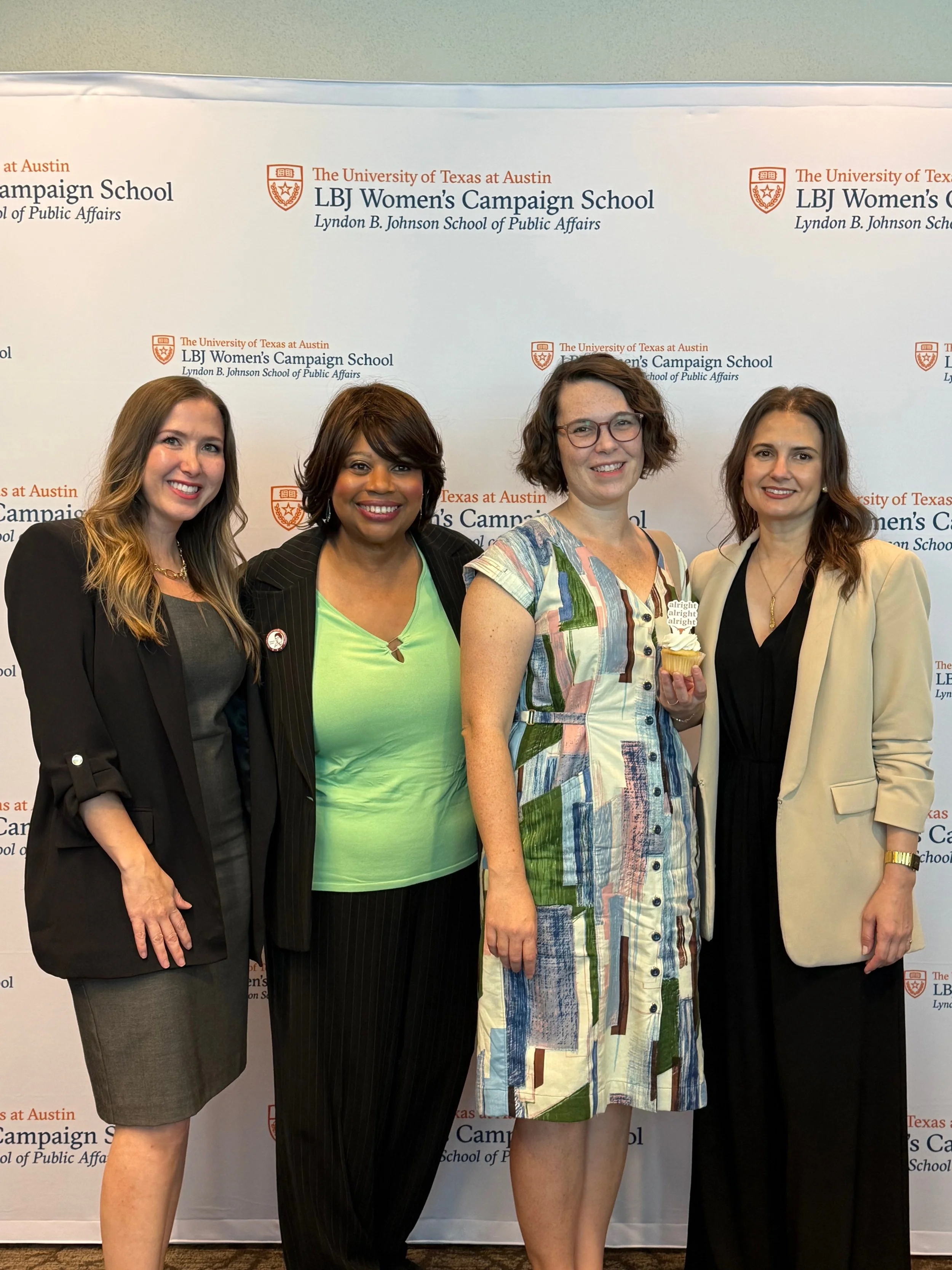 Four women standing together in front of a backdrop with the logos of the LBJ Women's Campaign School at the University of Texas at Austin, smiling for the camera. One woman is holding a cupcake.