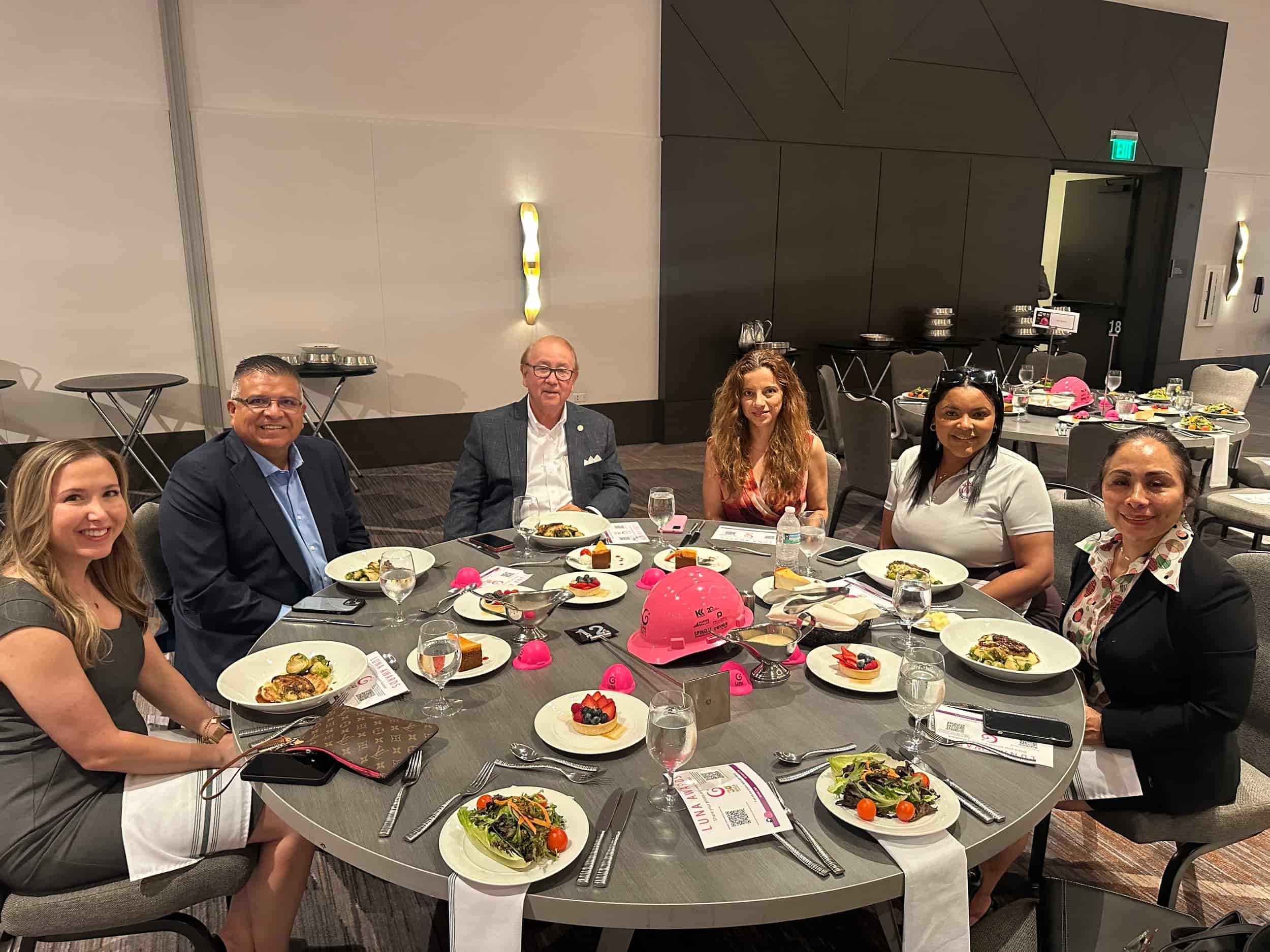 Group of six people seated around a dinner table at a formal event, with plates of salad, dessert, and drinks, in a modern banquet hall.