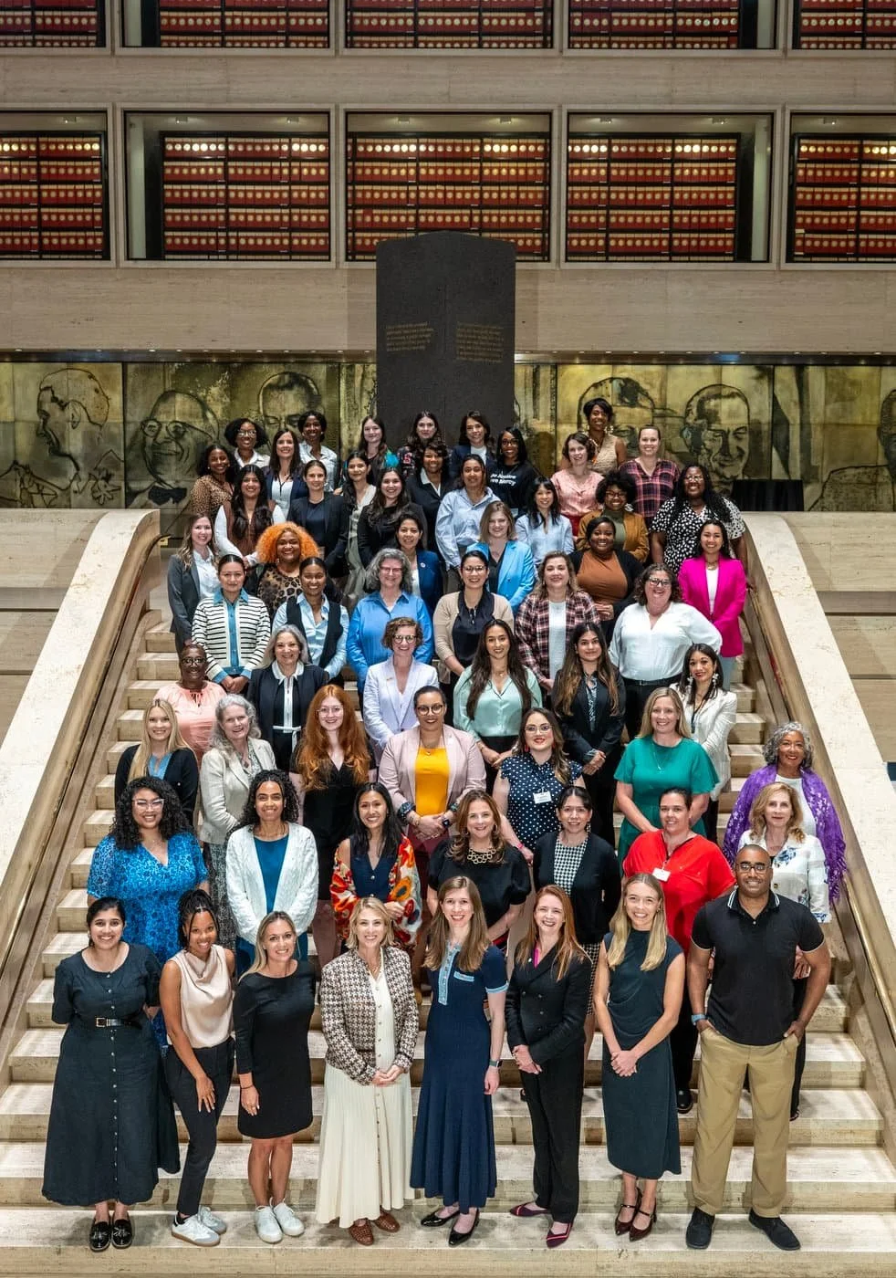 A large group of women and a few men standing on a staircase inside a building, likely at a professional or academic event, with a memorial or art installation on the wall behind them.
