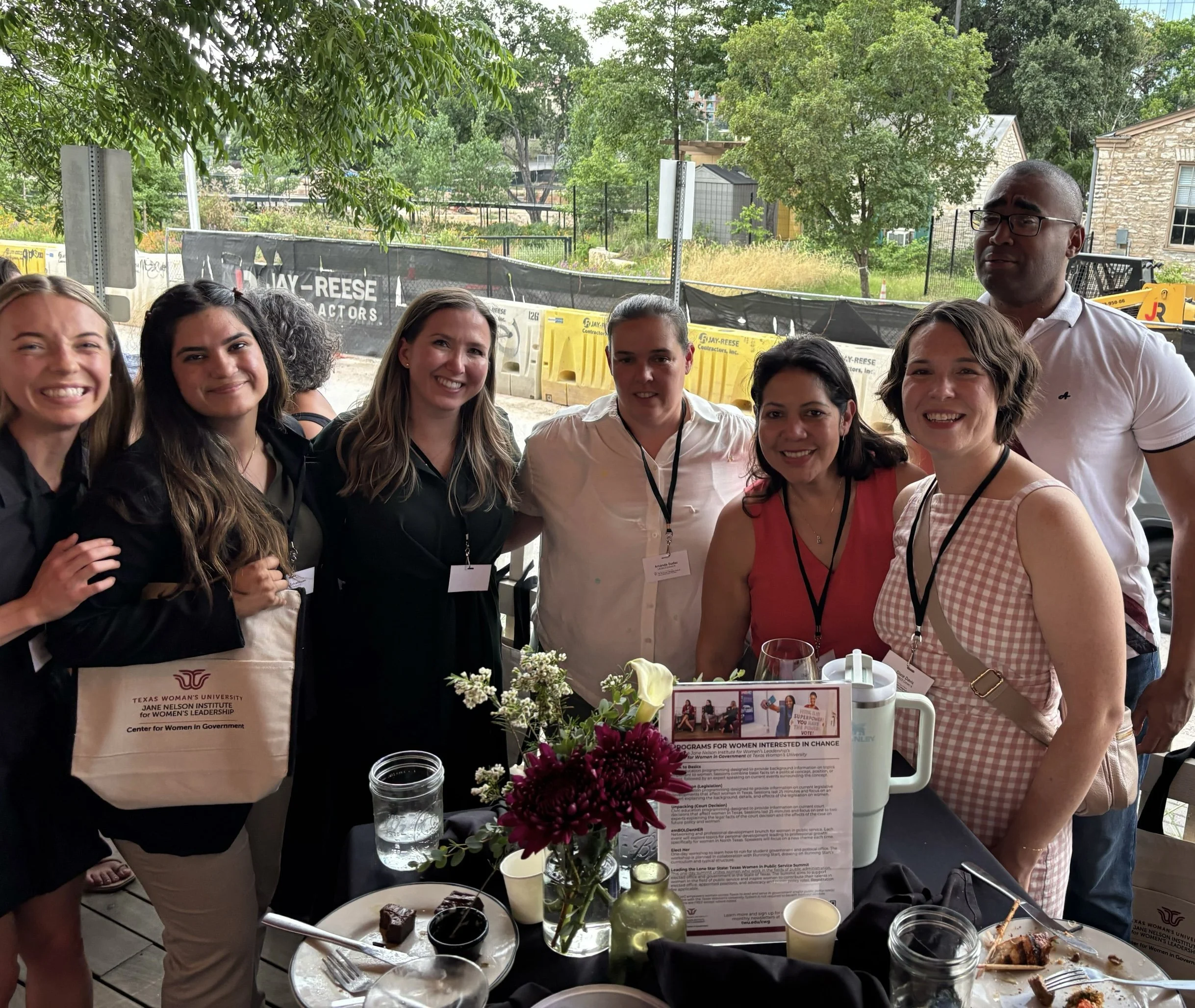 Group of seven diverse women and one man smiling at an outdoor event, standing around a table with flowers and food, with construction and trees in the background.