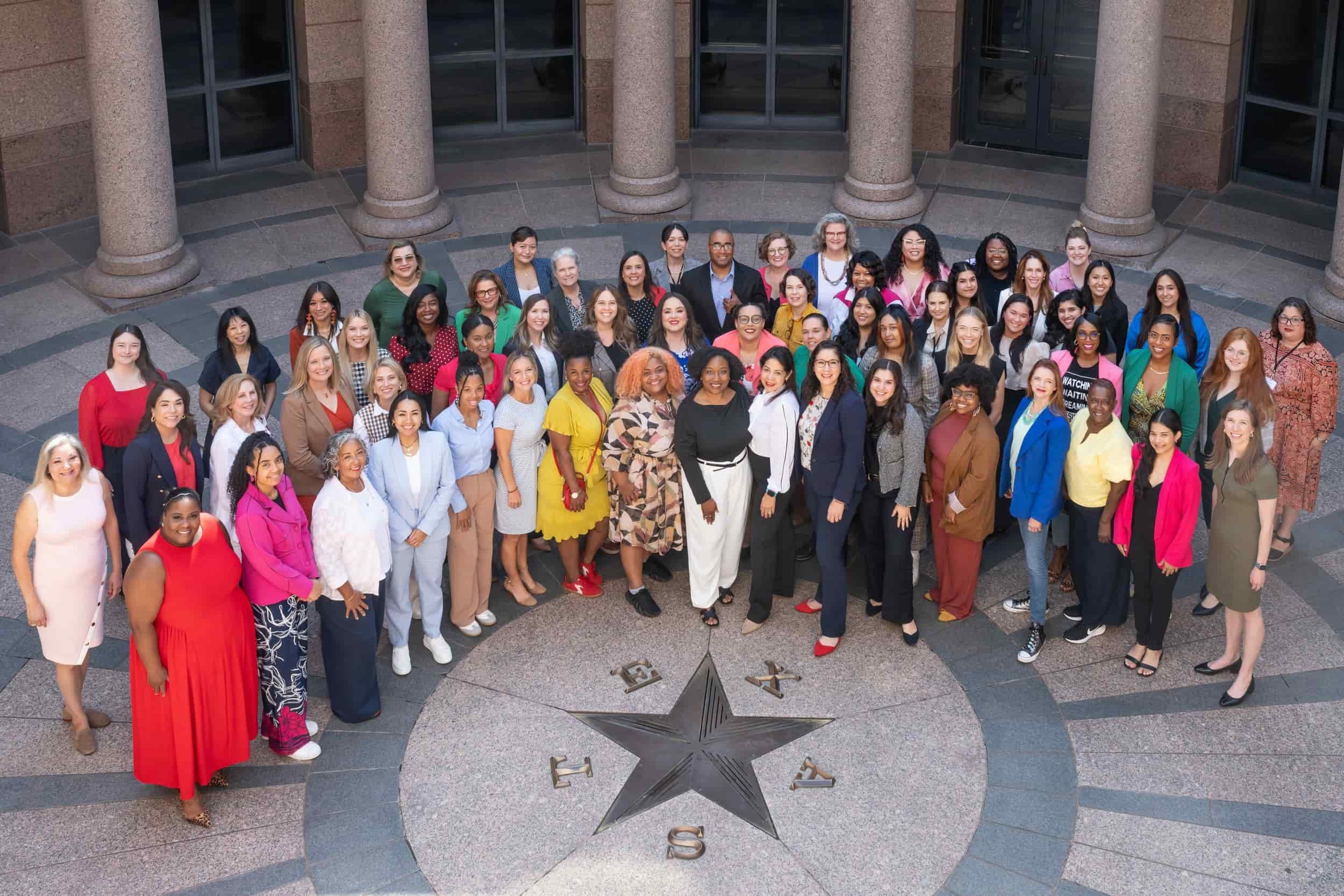 A large group of diverse women and men gathered in a semi-circle around a star emblem on the ground, located outdoors in front of a building with tall columns and glass windows.