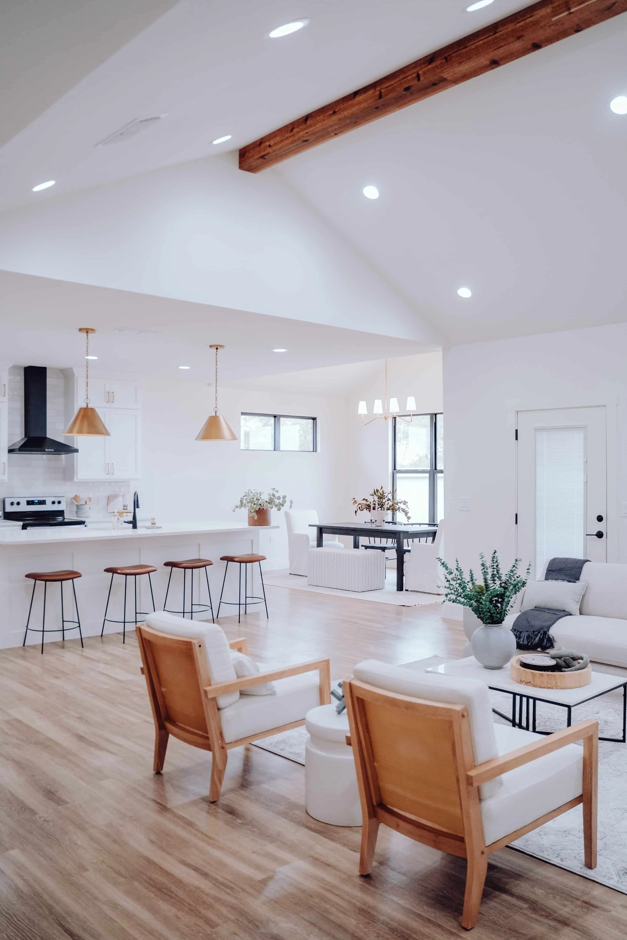 Bright white kitchen with three woven pendant lights, white cabinets, a stainless steel range hood, a small wine cooler, and a kitchen island with wicker chairs. Decor includes fresh green artichokes, potted plants, and a large vase with greenery.