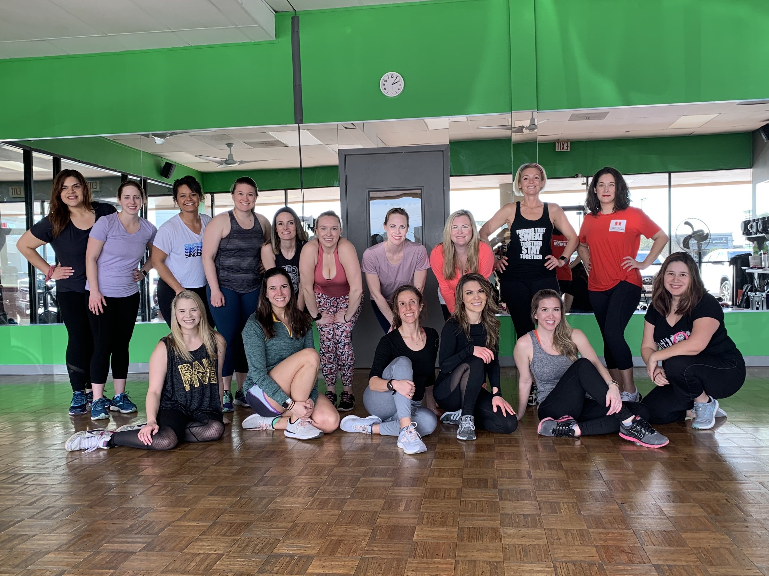 Group of women in workout clothes posing in a fitness studio with green walls, wooden floor, large mirror, and windows in the background.