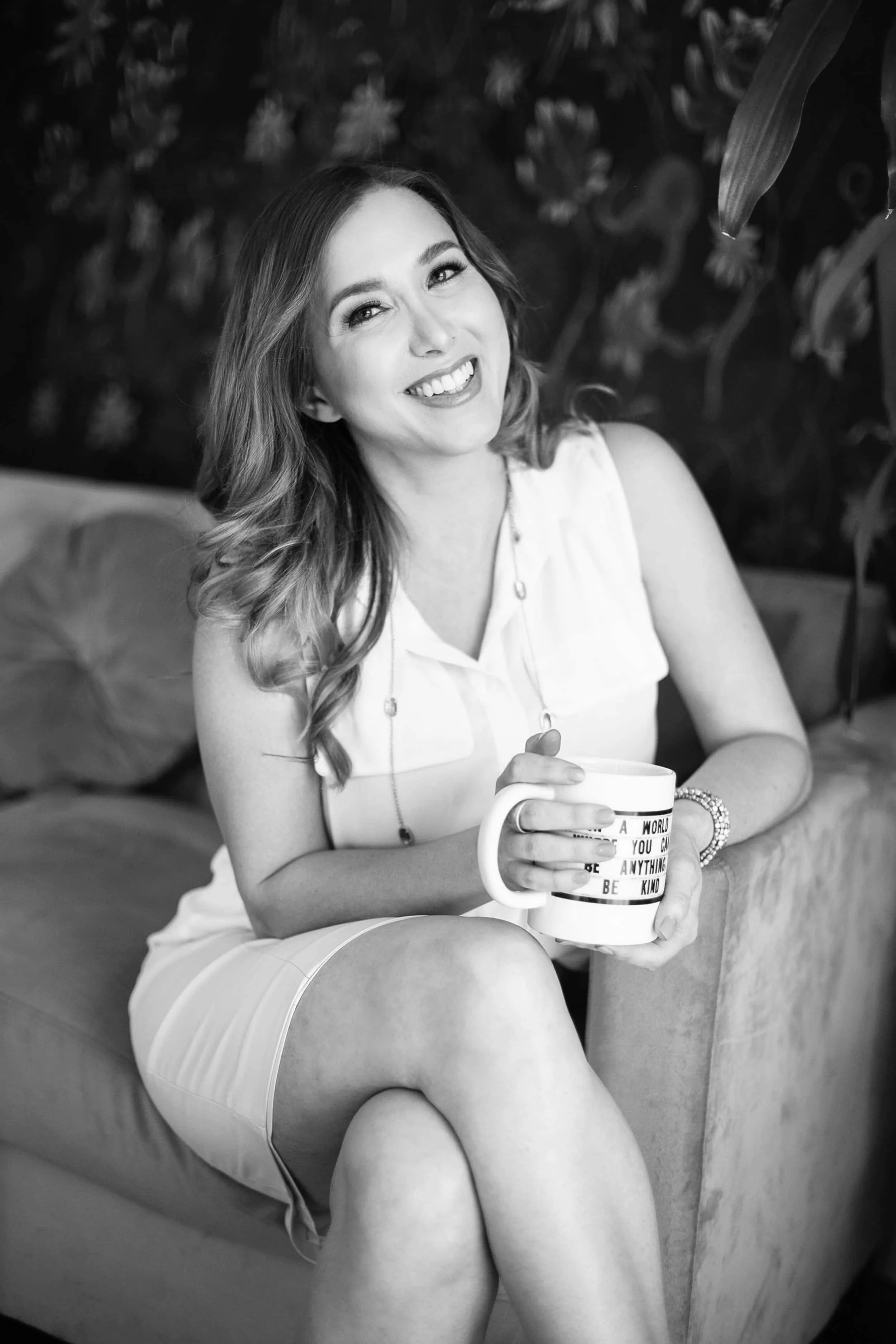 April Guerra, a woman with light skin, wavy hair, and a bright smile sitting on a sofa, holding a mug with a motivational quote, in a setting with plants and a dark background.