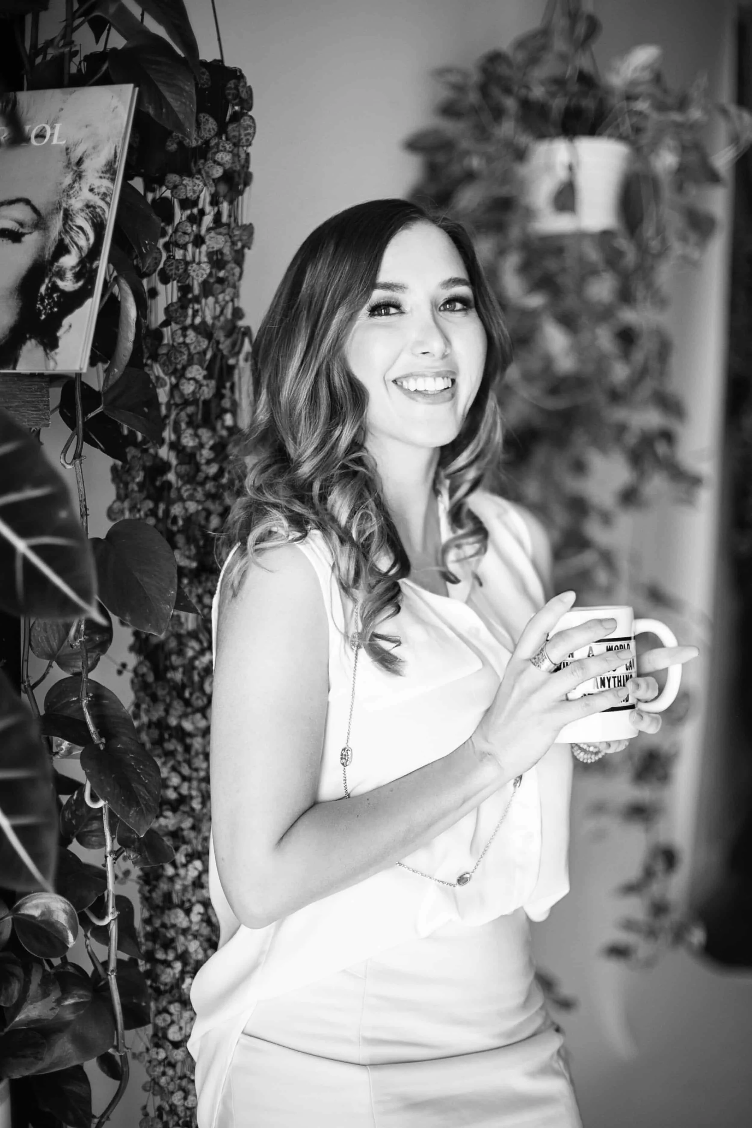 April Guerra, a woman with wavy hair smiling and holding a mug, standing next to plants and a Marilyn Monroe poster.