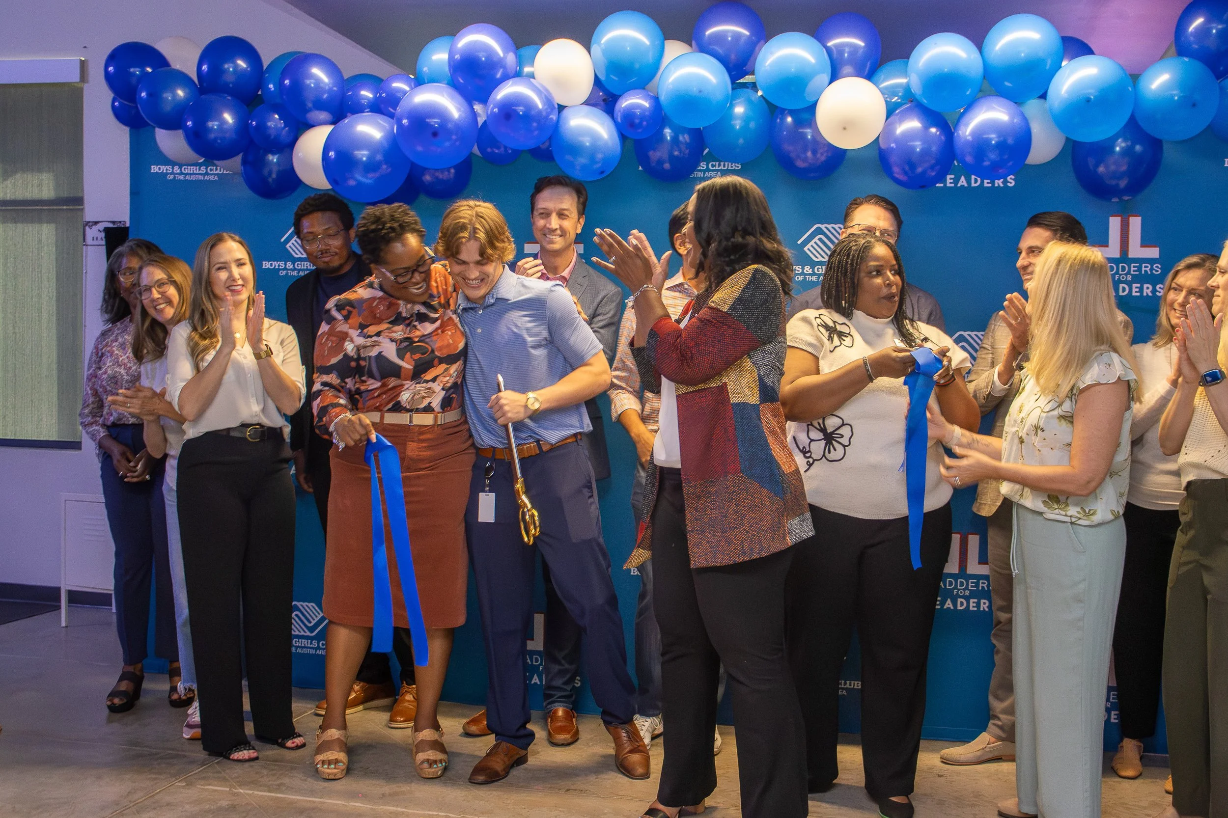 A diverse group of people at an event, smiling, talking, and holding ribbons, with blue and white balloons in the background at the Boys & Girls Clubs of the Austin Area.