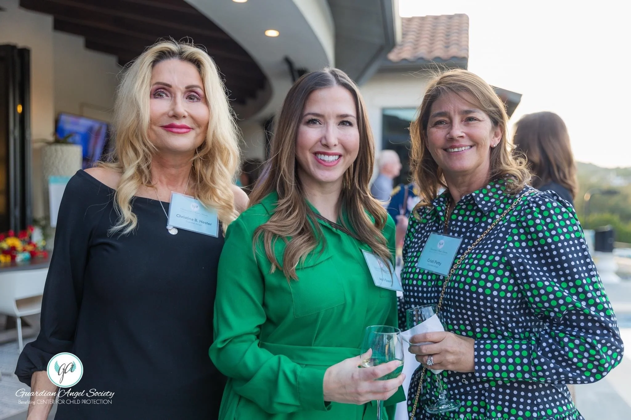 Three women at an outdoor event, smiling and holding drinks, wearing name tags, with a background of a building and trees.
