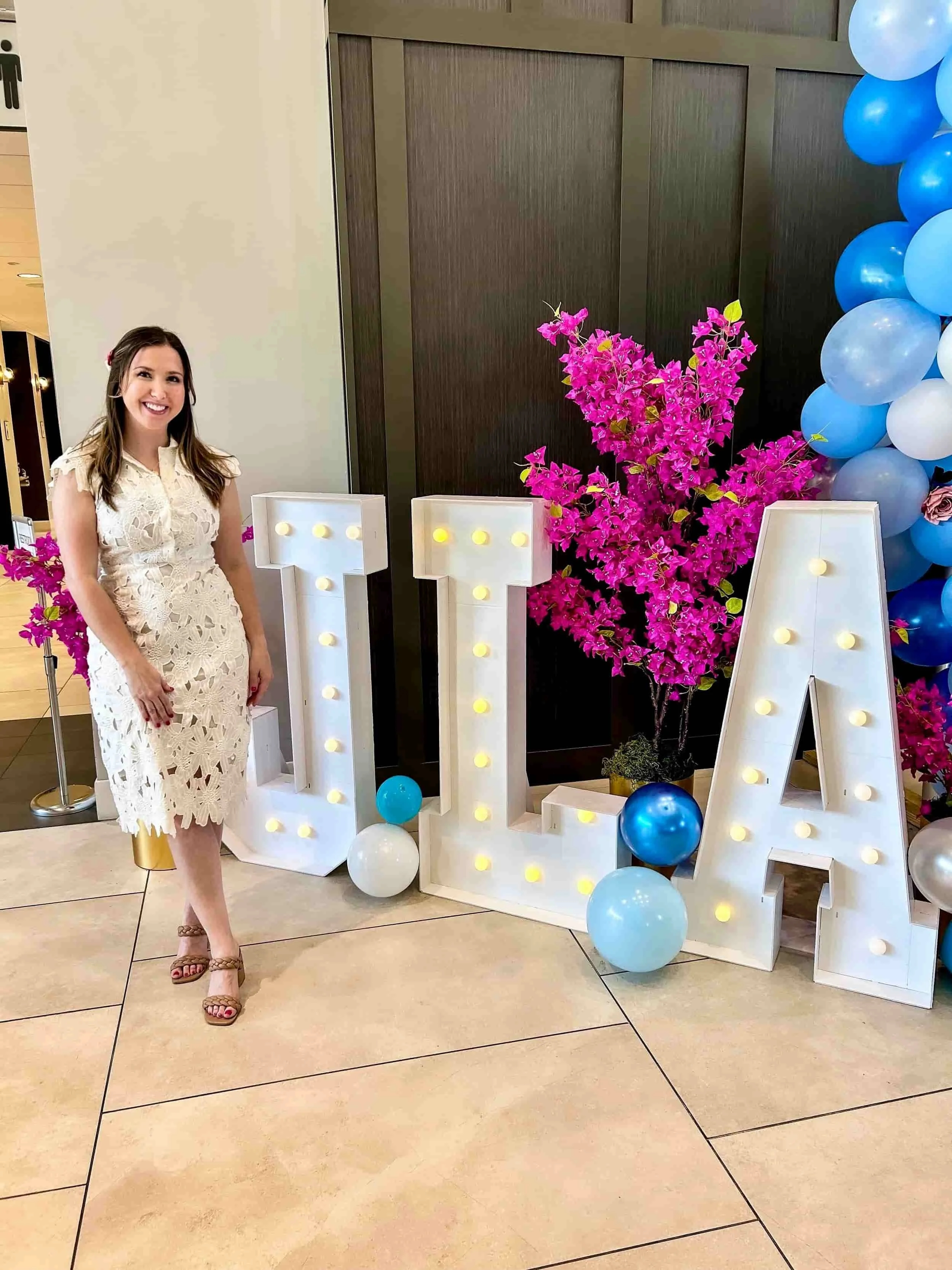 April Guerra in a white lace dress standing next to large marquee letters spelling 'JLA' with decorative balloons and pink flowers in the background.