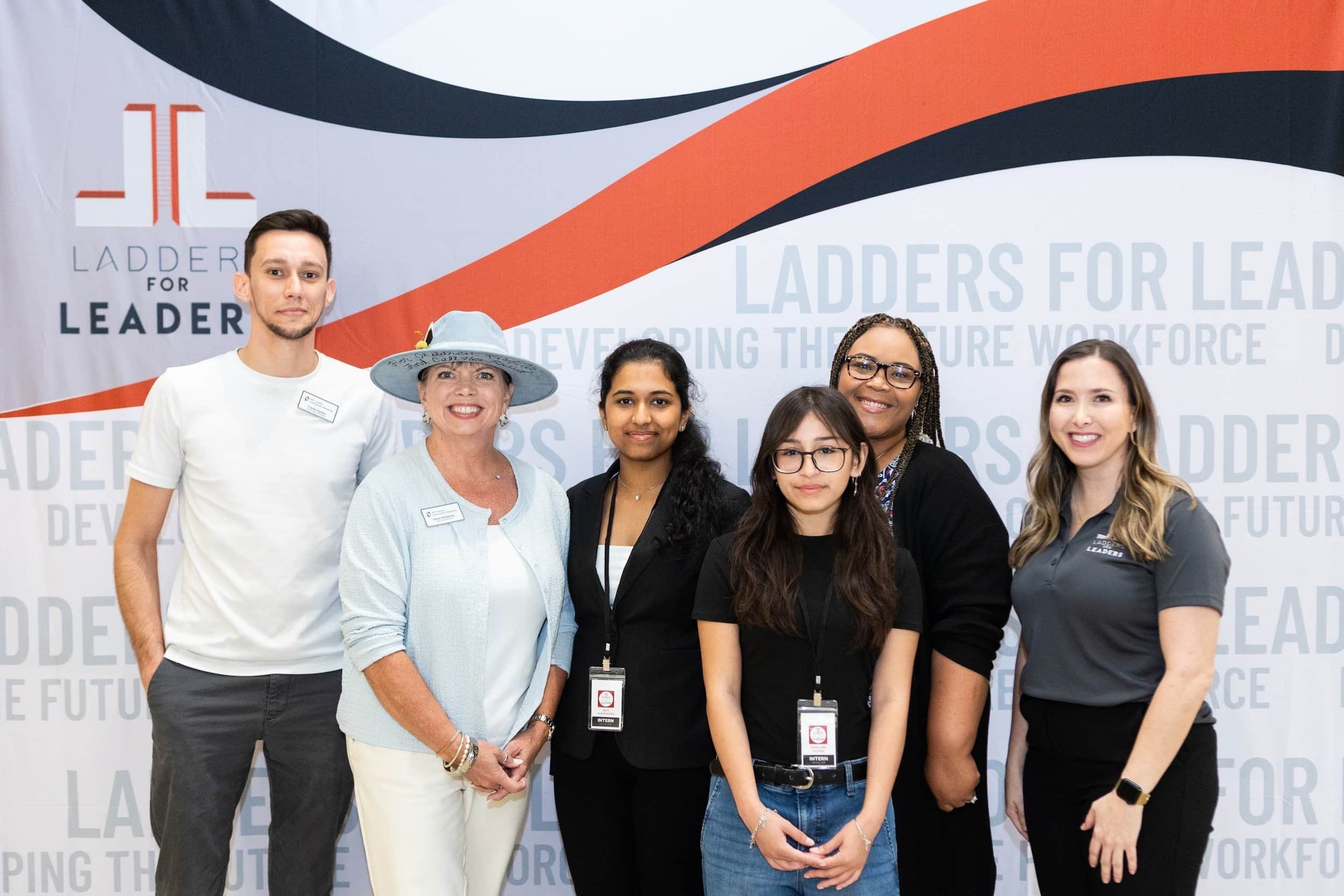 Group of six diverse individuals standing in front of a backdrop with the text 'LADDERS FOR LEADERS' and developing the future workforce' at a professional event, smiling at the camera.