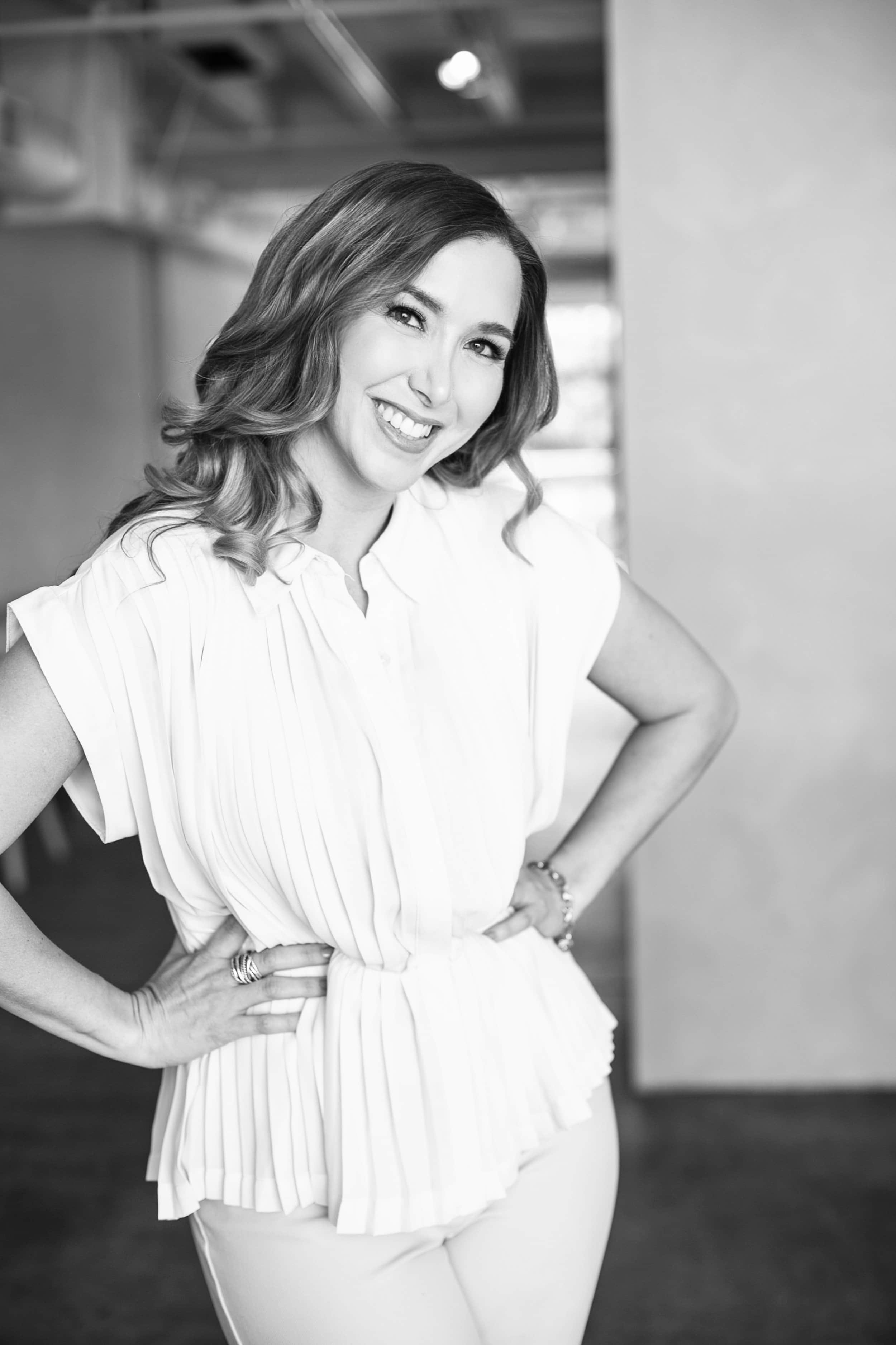 April Guerra, a smiling woman with wavy hair, wearing a light-colored pleated blouse and pants, standing indoors with hands on hips.