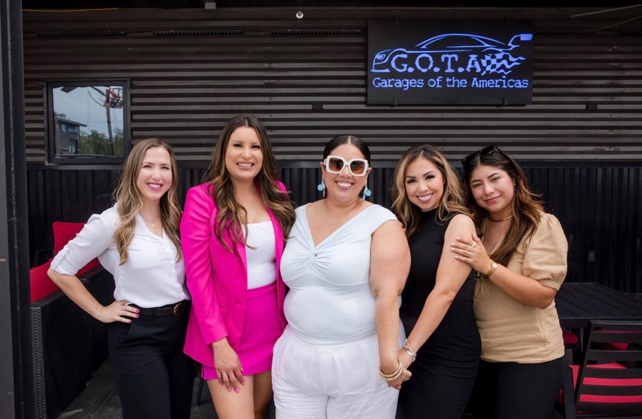 Five women smiling and standing together in front of a sign that reads 'G.O.T.A.W. Garages of the Americas,' outside a building with dark wood siding.