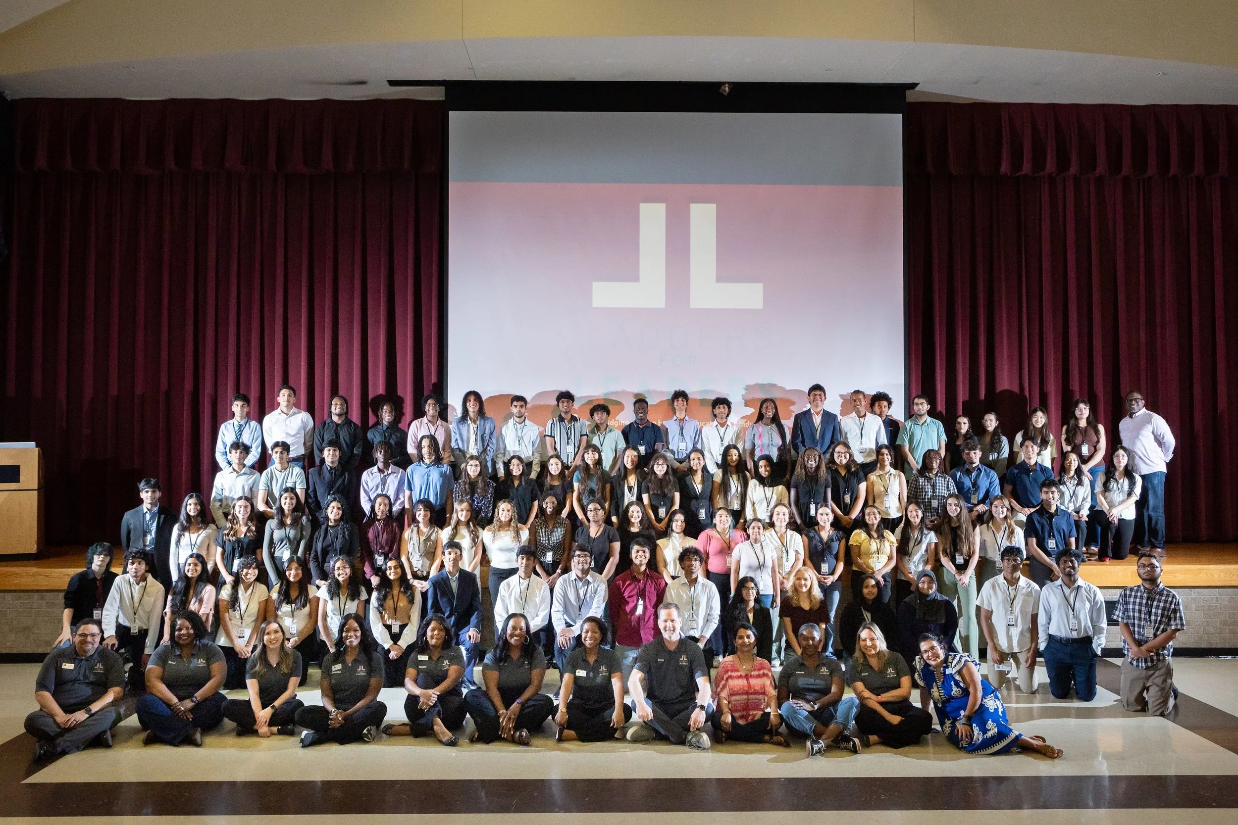 Group photo of diverse students and staff on stage during a school event, with a large screen displaying a logo in the background.