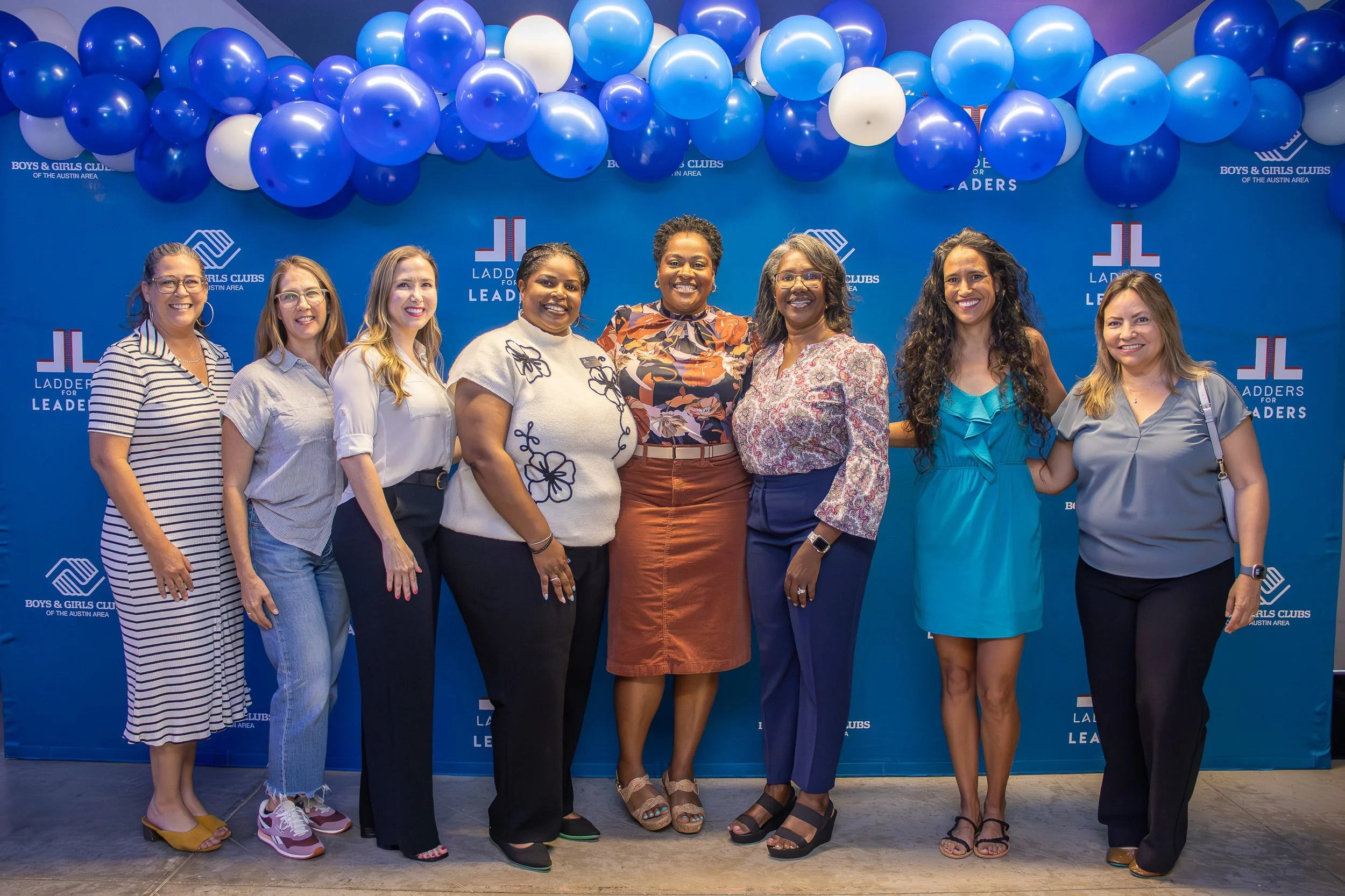 A group of eight women standing in front of a blue backdrop with the Boys & Girls Clubs of the Austin Area logo, under a large arrangement of blue and white balloons, smiling.