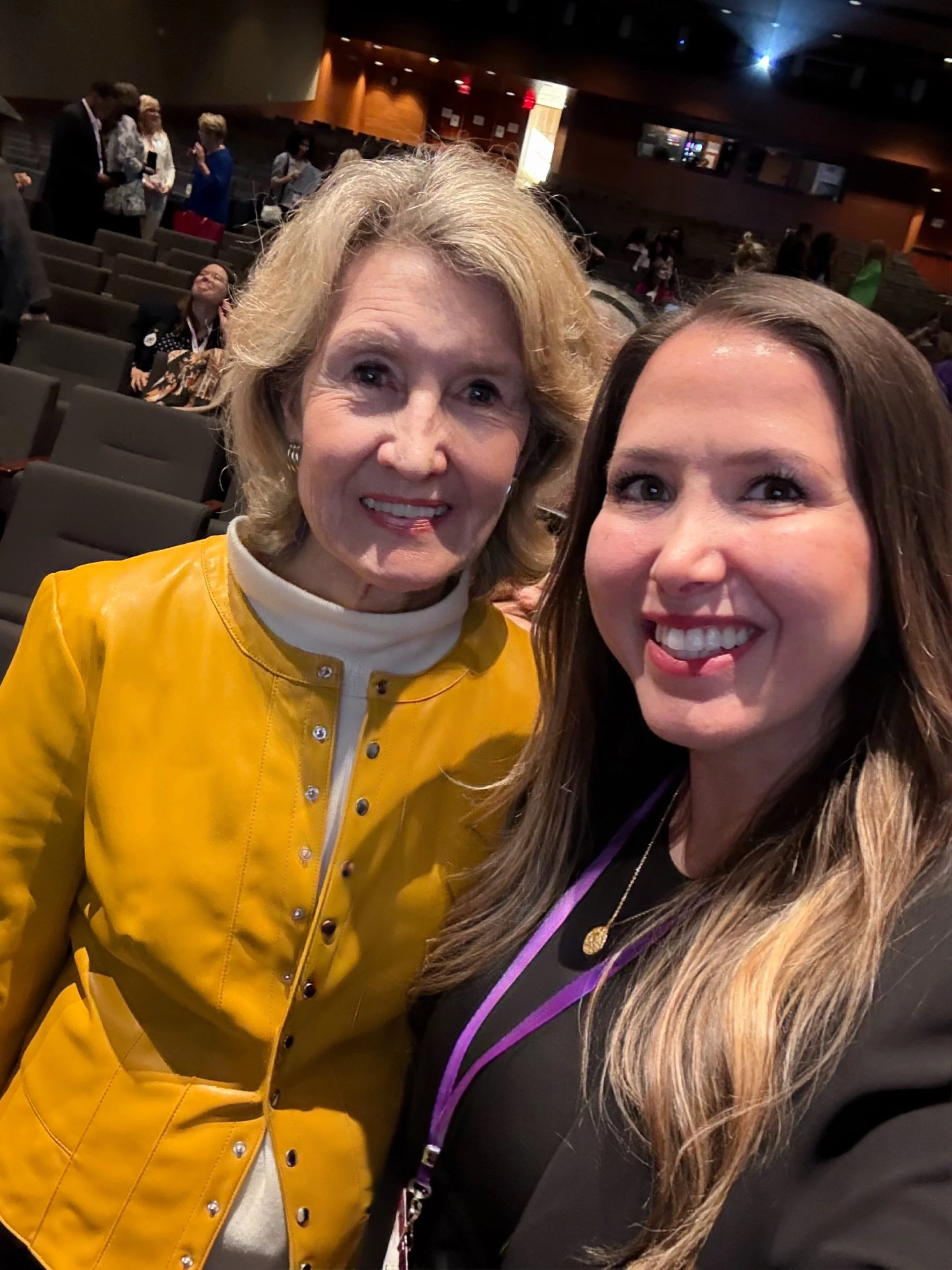 Two women smiling for a selfie in an auditorium, with other people in the background.