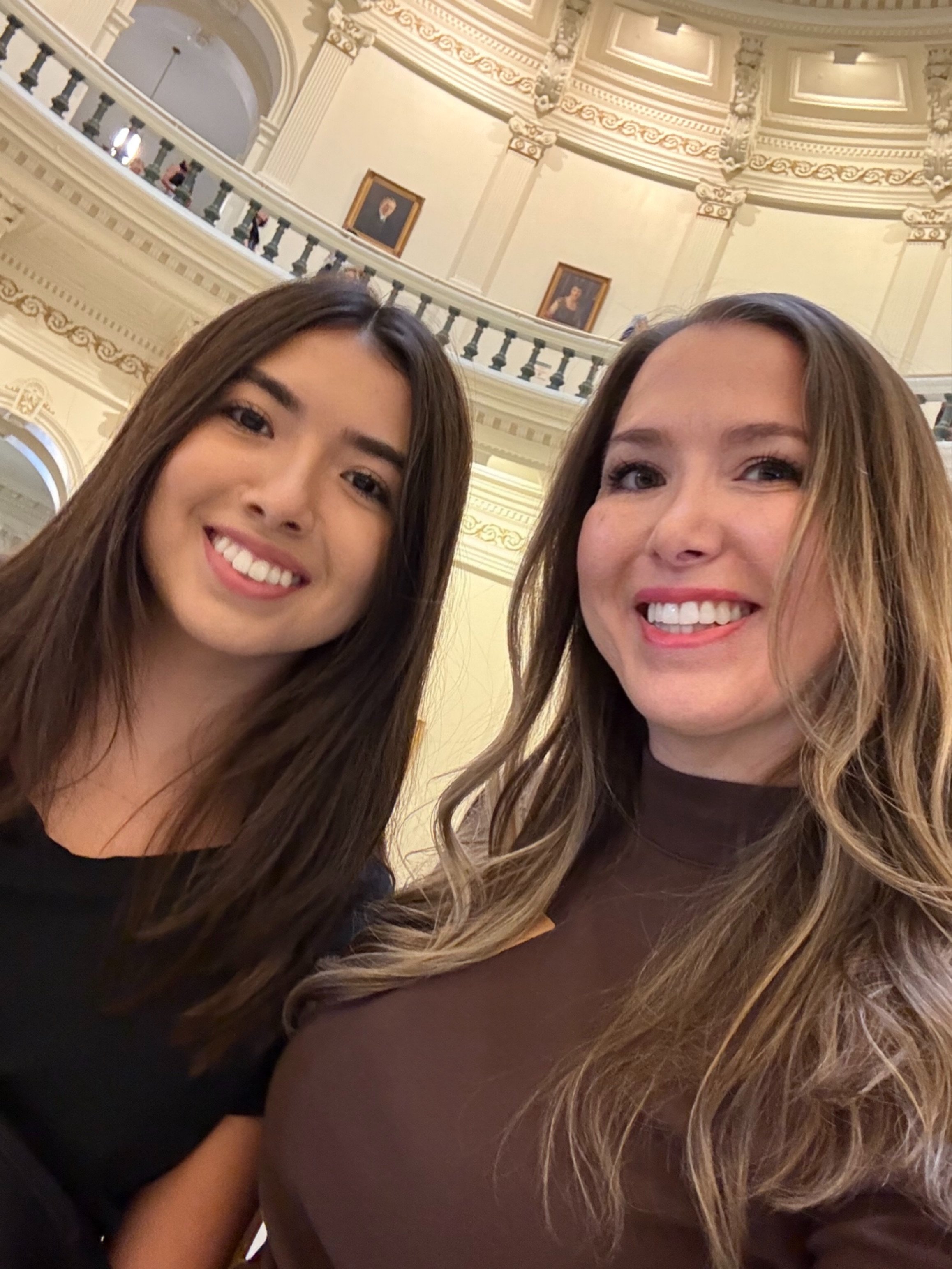 Two women smiling for a selfie inside a grand, ornate building with a curved balcony, detailed molding, and portraits on the upper wall.