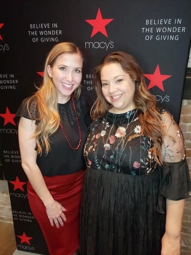 Two women smiling and posing in front of a Macy's backdrop with the Macy's logo and the slogan 'Believe in the Wonder of Giving'.