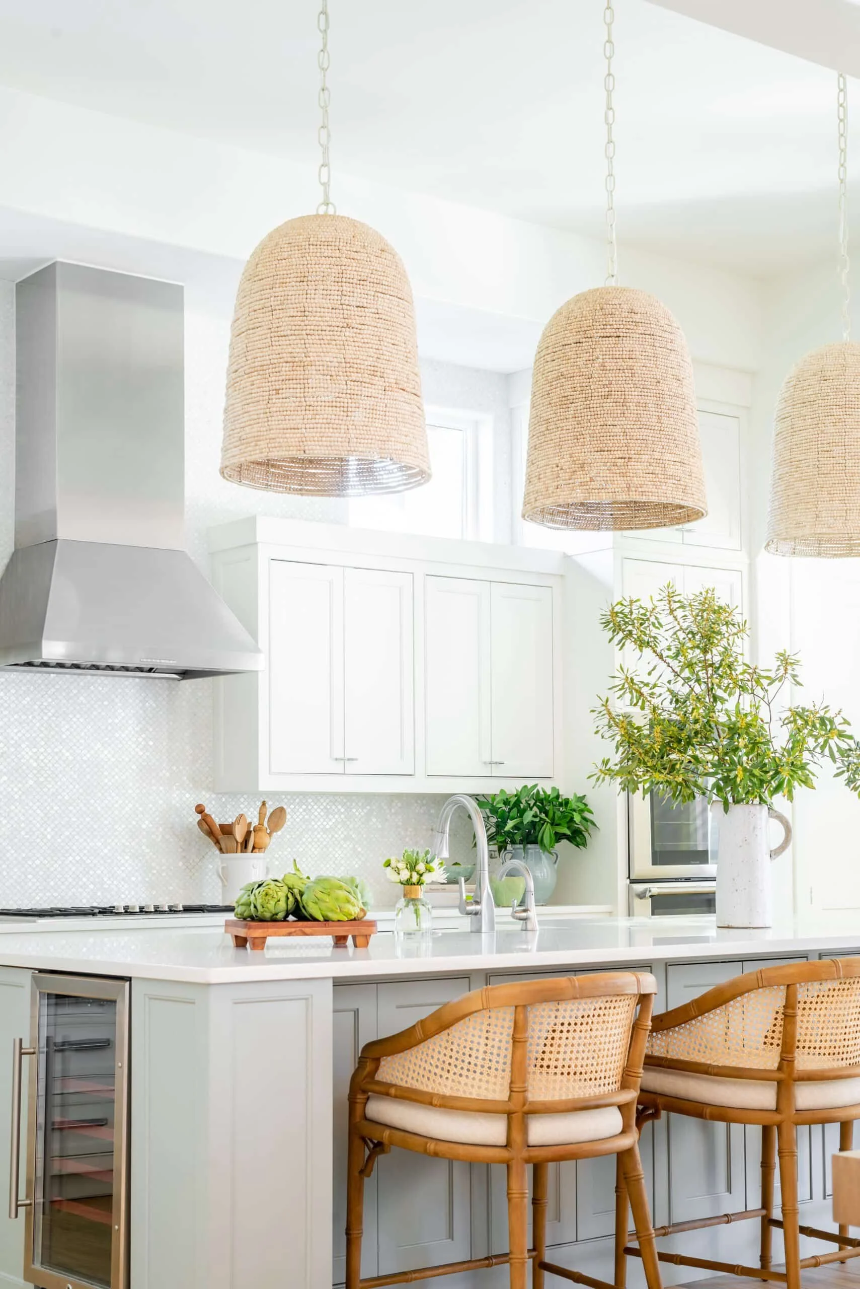 Modern kitchen with light wood cabinets and white marble countertop, gold pendant lights, white farmhouse sink, and green plant in a beige textured vase.