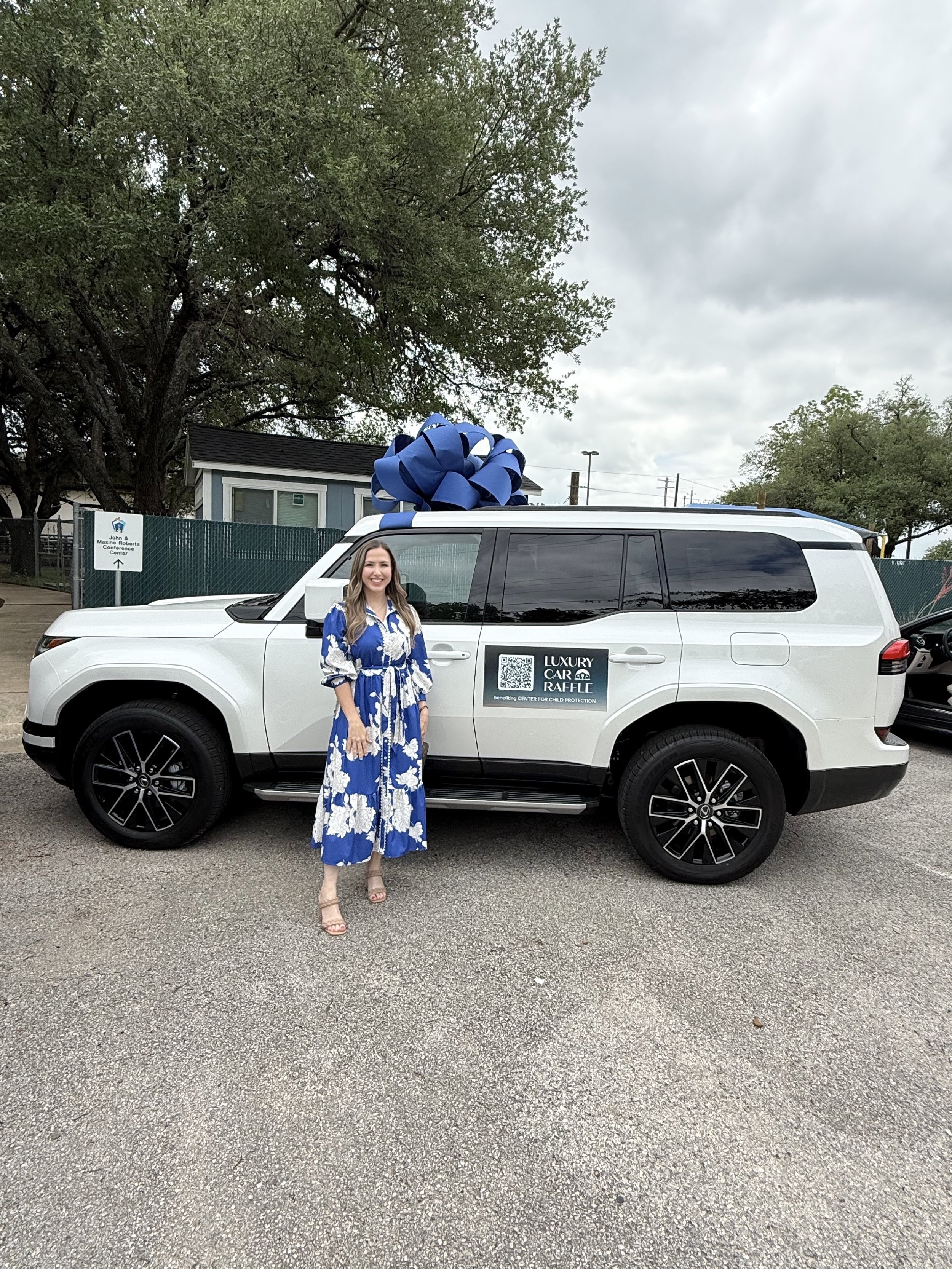 A woman wearing a blue and white floral dress standing next to a white vehicle with a large blue bow on the roof. The vehicle has a sign that reads 'Luxury Car Raffle benefiting Center for Child Protection.' The background includes trees, a small bui