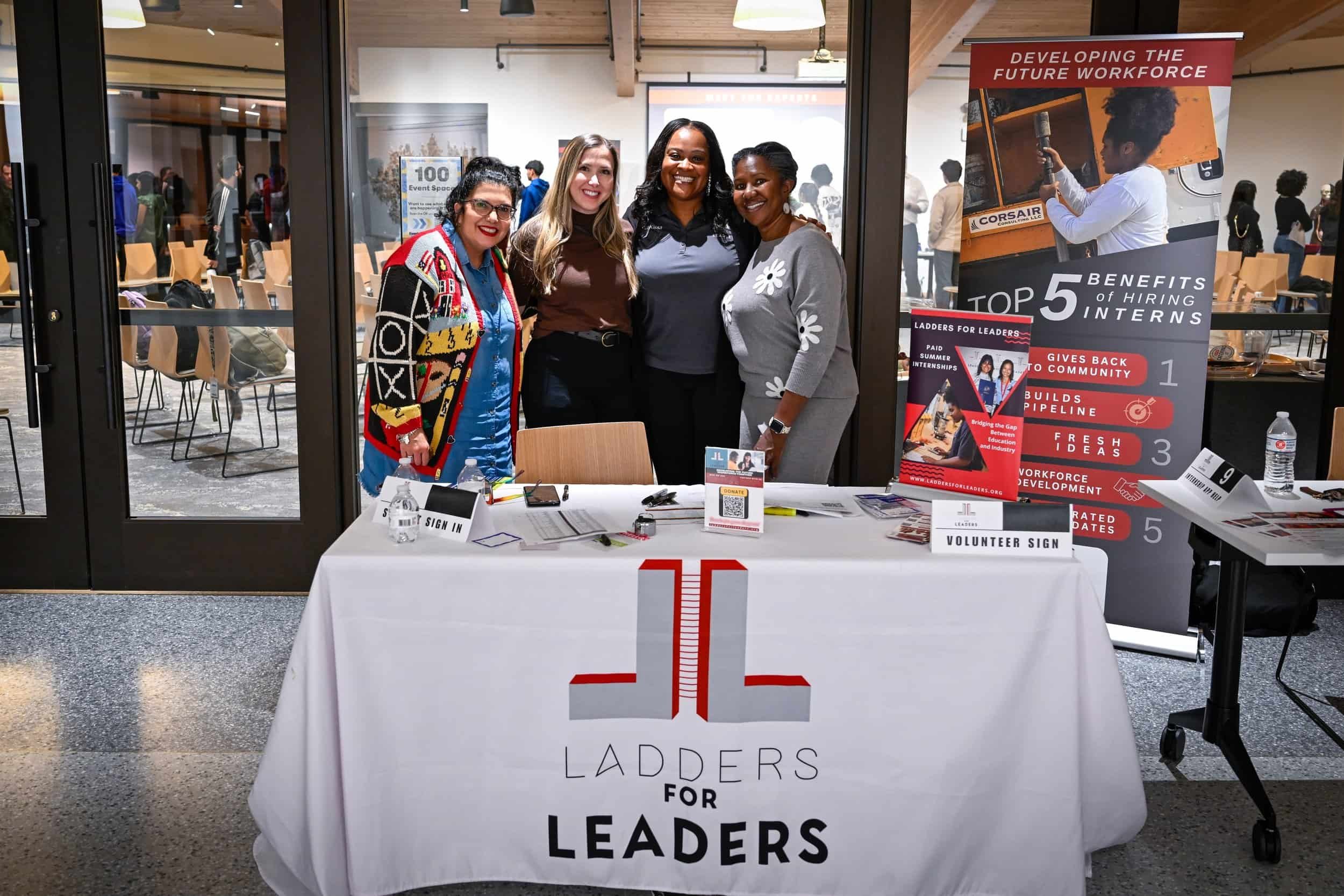 Four women standing behind a table at a career event, with signs and informational materials displayed, in front of a door leading to a conference or event space.
