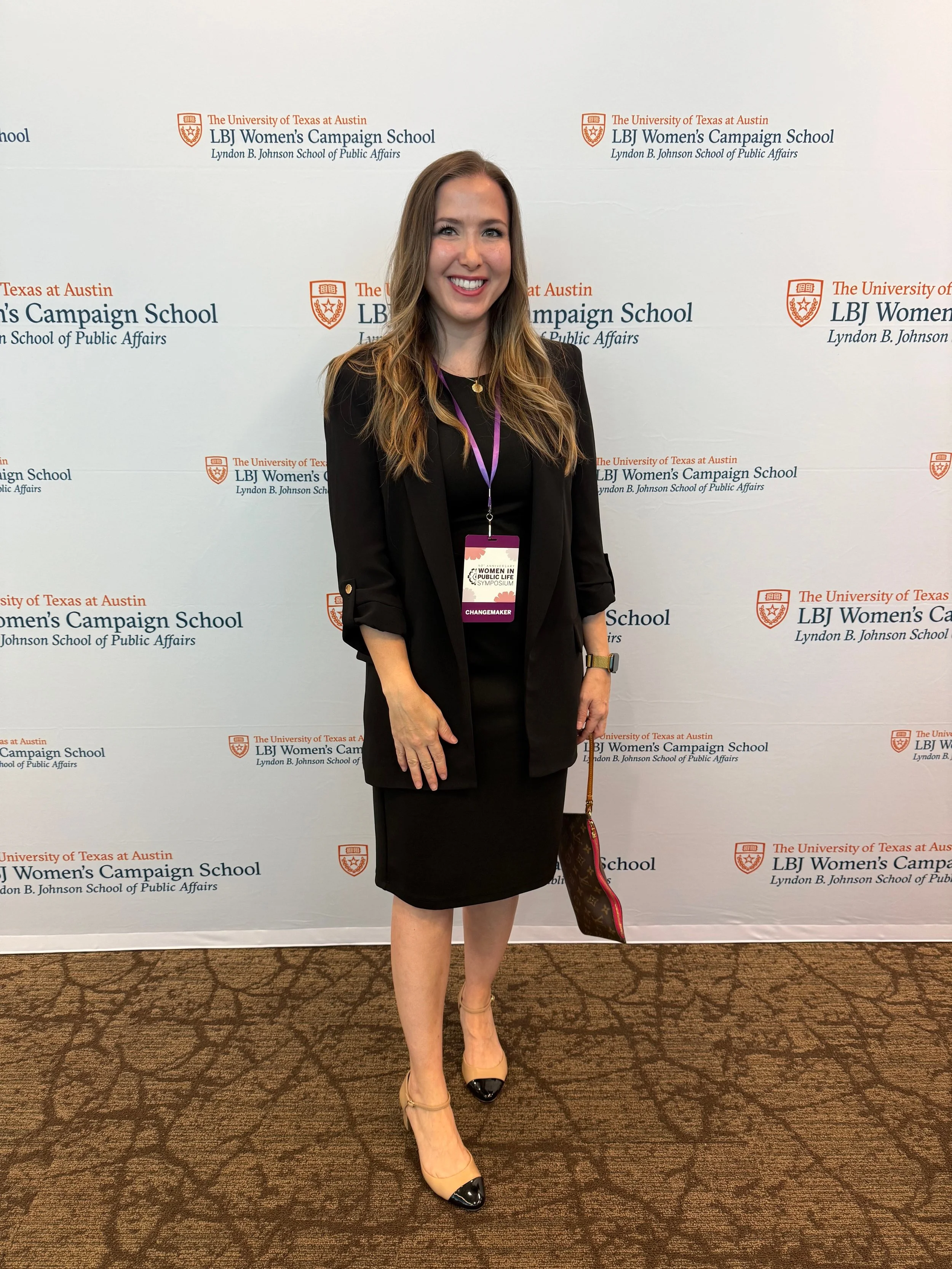 Woman smiling, wearing a black blazer and dress, standing in front of a backdrop with the LBJ Women's Campaign School and University of Texas at Austin logos, holding a purse and wearing a conference badge.