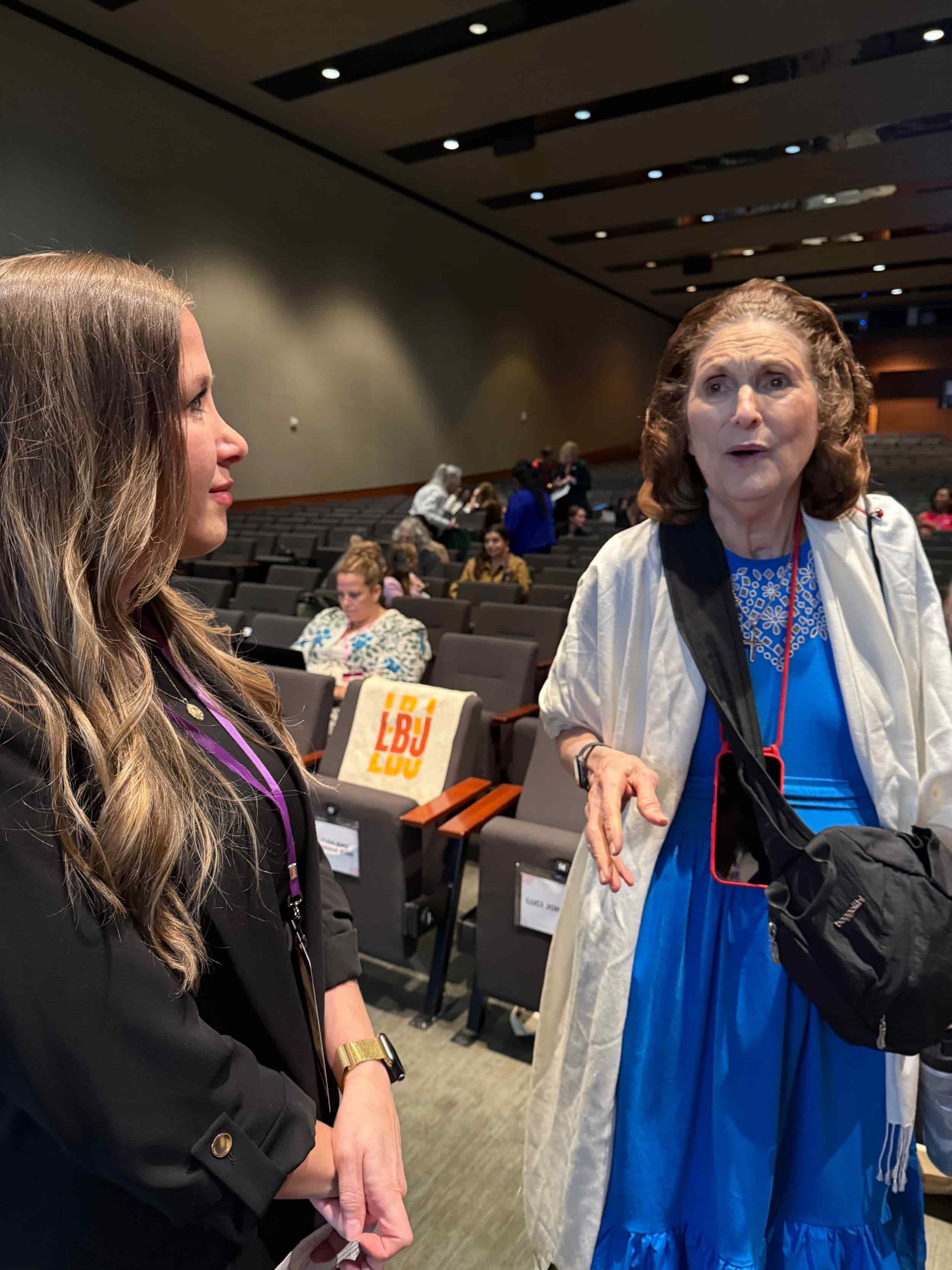 Two women talking in an auditorium, with several people seated in the background, including one with a tote bag with the logo 'LBU LDU'.