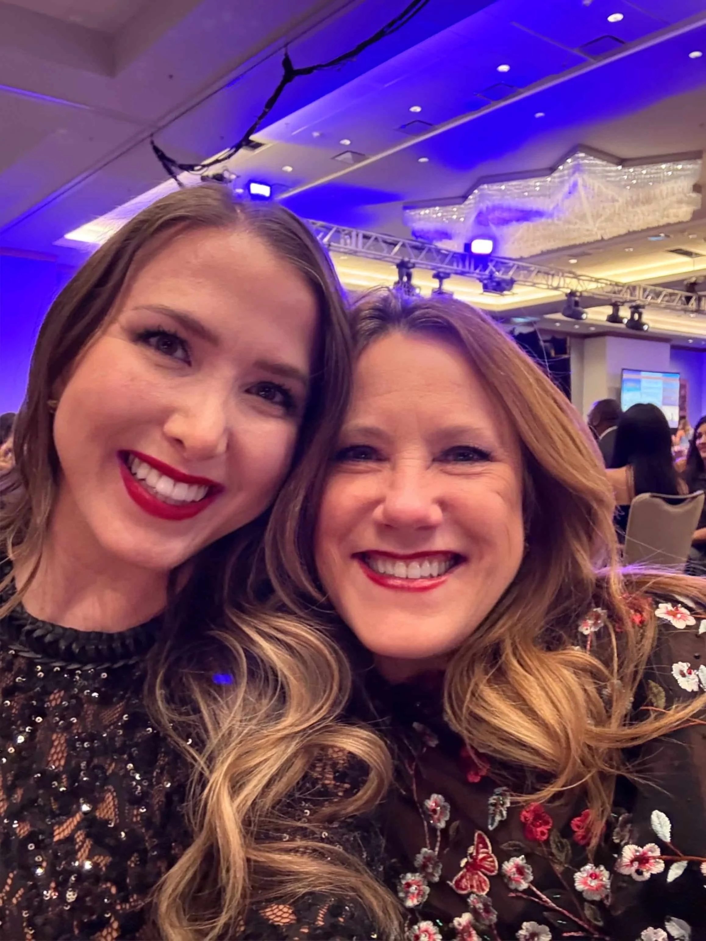 Two women smiling and taking a selfie at an event with blue lighting and a chandelier in the background.