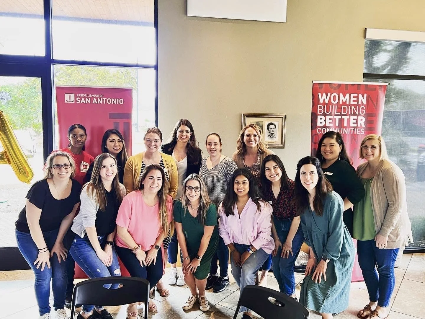 Group of diverse women smiling at a community event inside a building with banners that say 'Women Building Better Communities' and 'Junior League of San Antonio'.