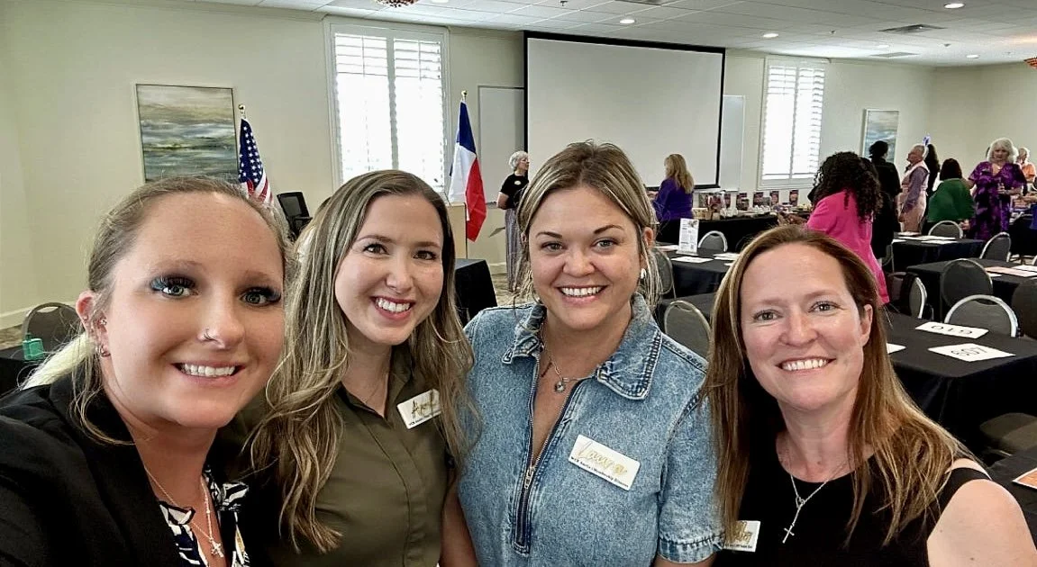 Four women smiling at a conference or meeting room with tables, chairs, American and Texas flags, and a projection screen in the background.